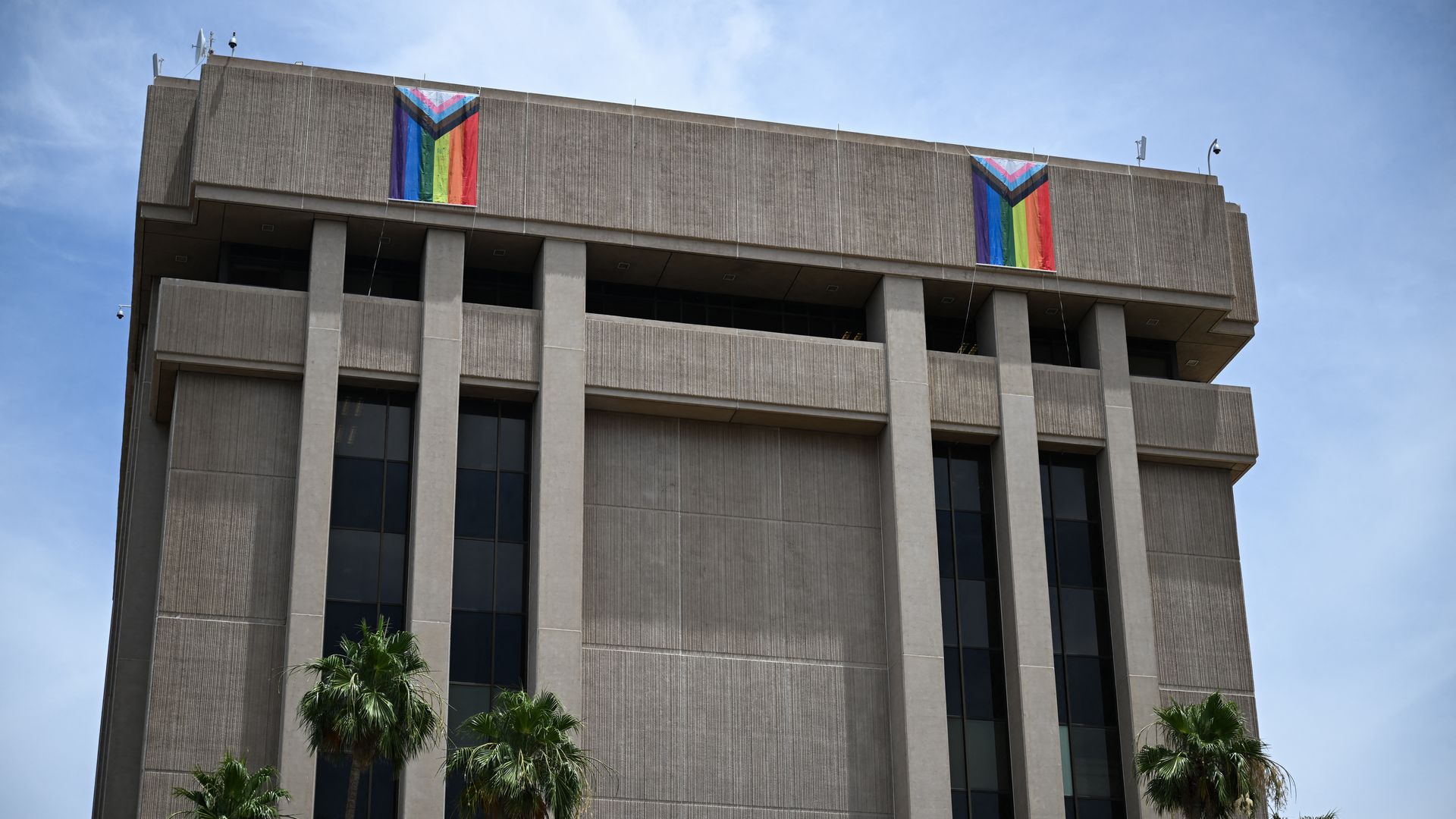 Tall concrete office building with two large rainbow Pride banners on the upper facade; clear blue sky and palm trees at the base.