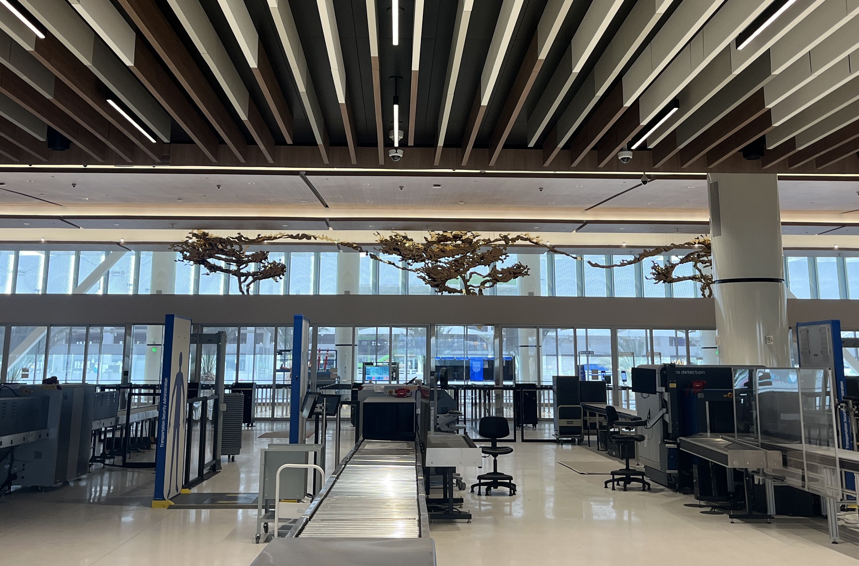 Interior view of a modern airport security checkpoint with metal detectors, conveyor belts, black chairs, and large windows showing an outside area, under a ceiling with wooden beams.