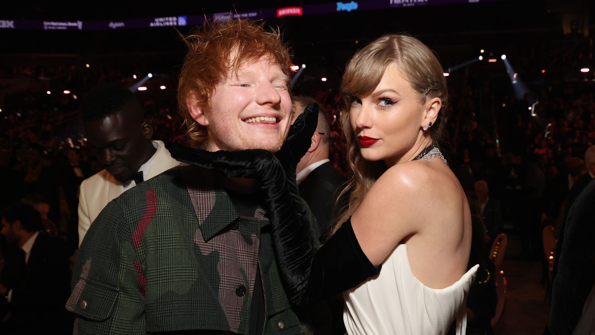 Taylor Swift dressed in evening wear places a gloved hand under Ed Sheeran's chin while looking over her shoulder at the camera, while Sheeran smiles.