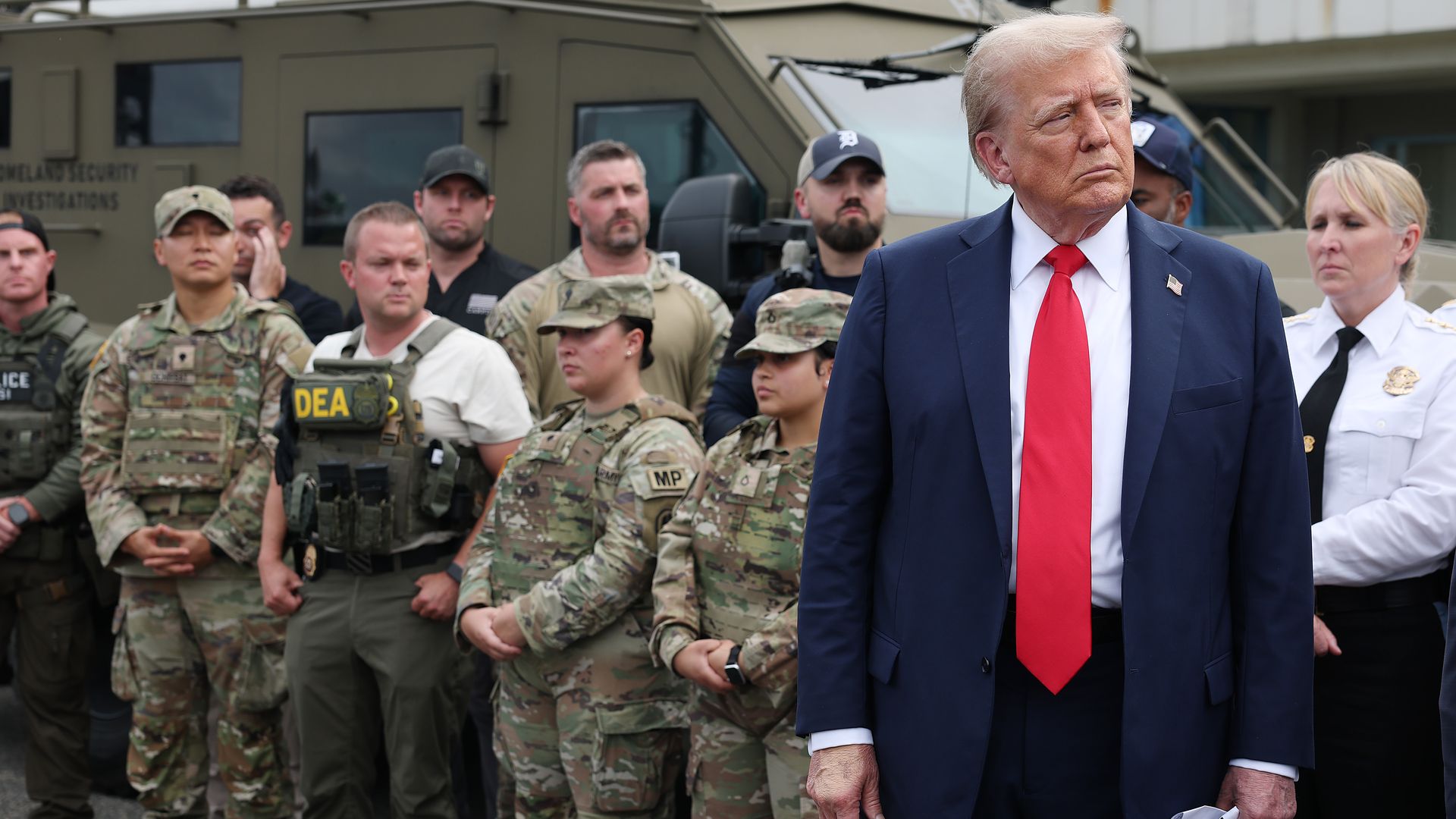 Man in navy suit with red tie holding white cap stands next to group of uniformed military and law enforcement personnel in front of a tactical vehicle.