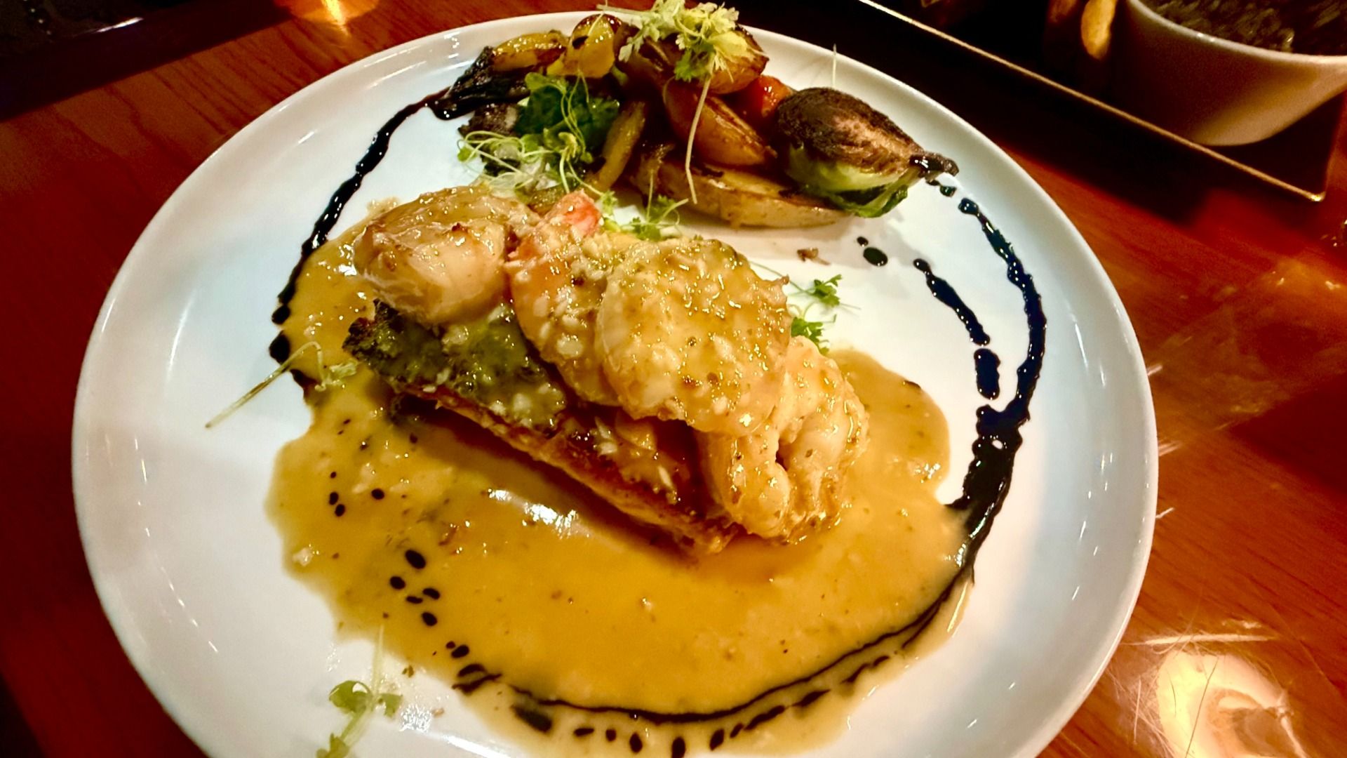 White plate with shrimp on a piece of fish topped with creamy yellow sauce, drizzled with dark balsamic glaze, served with roasted vegetables and greens on a wooden table.