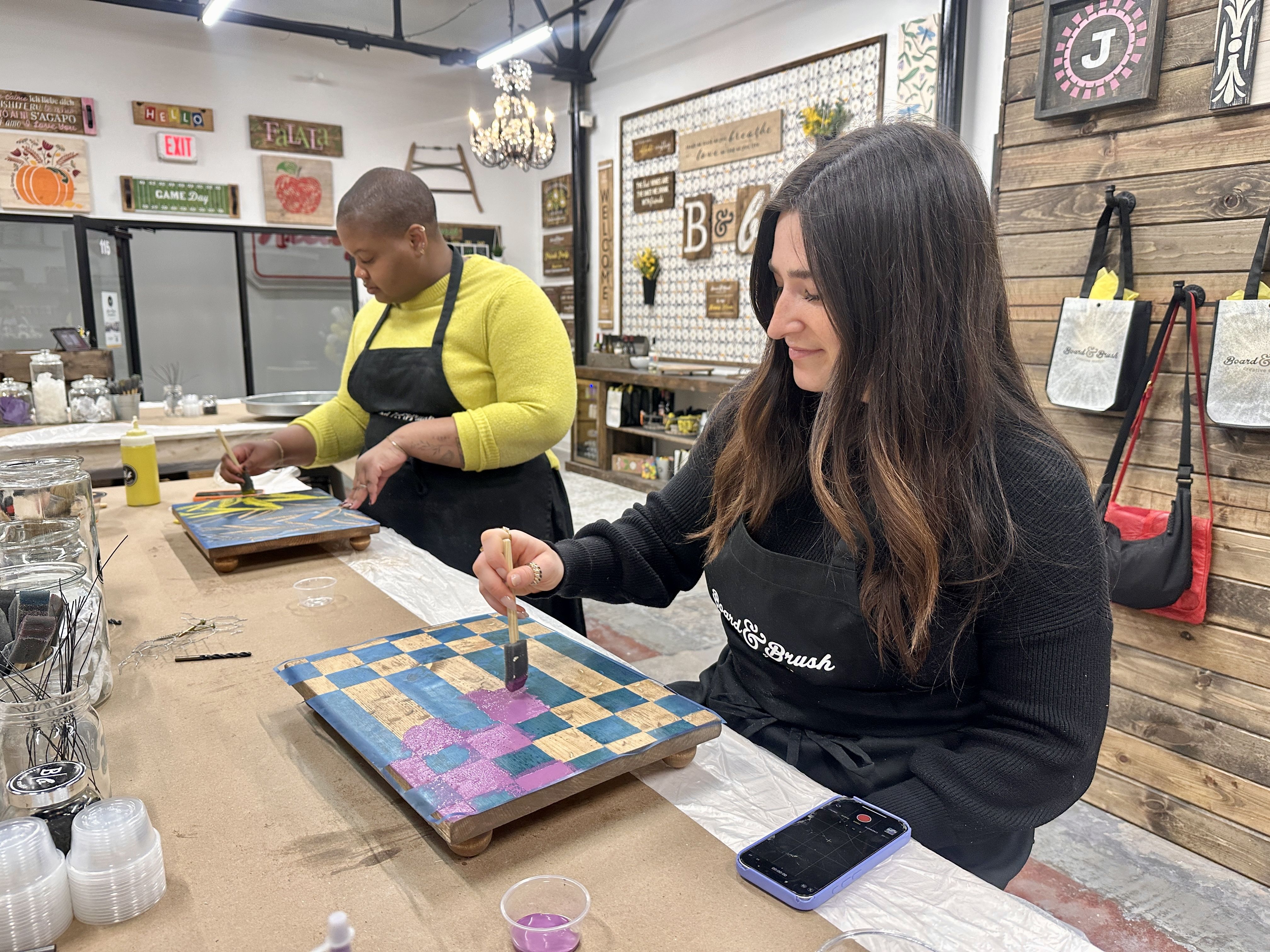 Two people paint wooden boards at a craft studio. A woman with long dark hair in a black apron paints a blue-and-tan checkerboard board; in the background, a man in a yellow sweater works on another board.