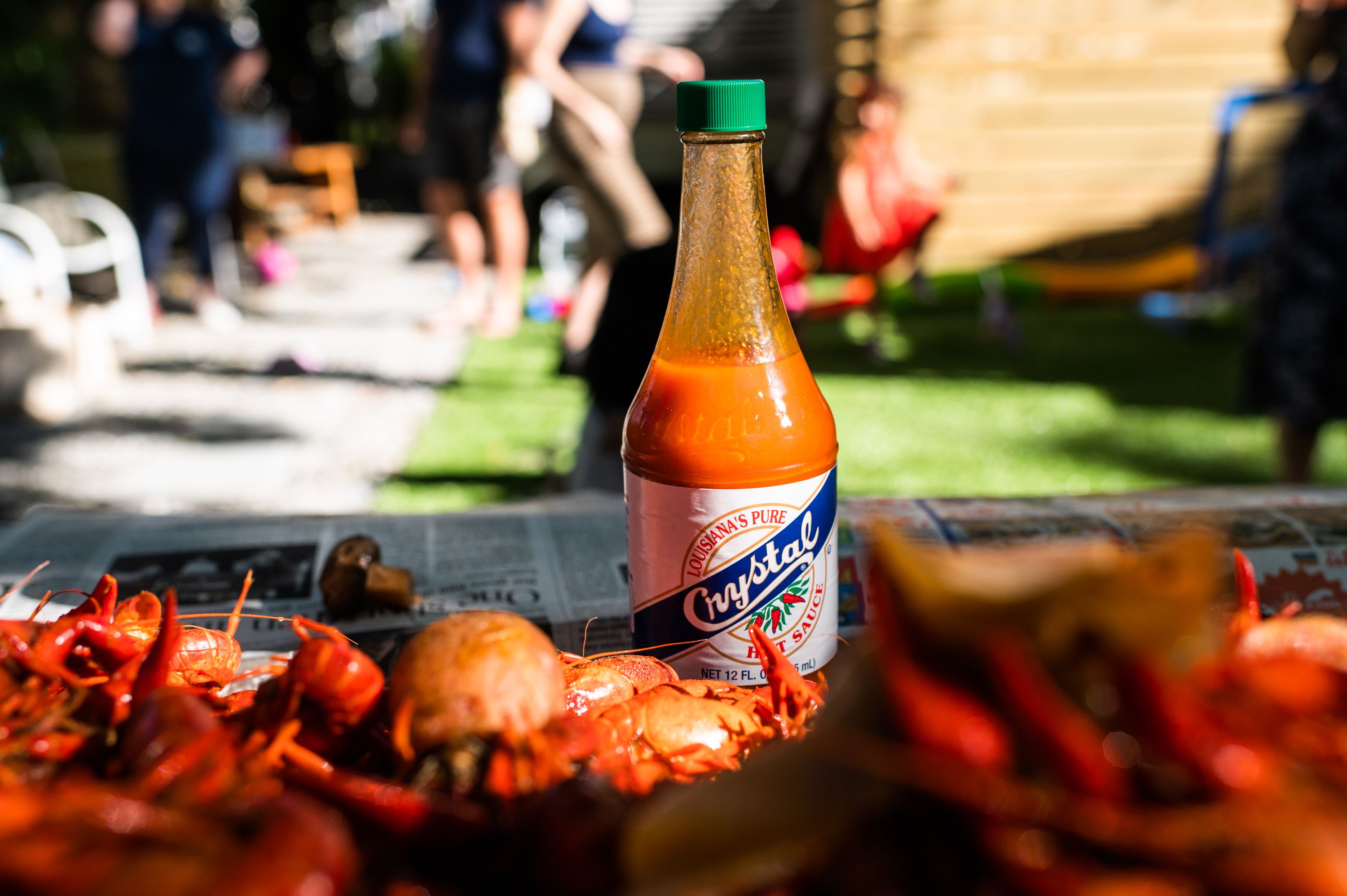 A bottle of Crystal hot sauce with a green cap is surrounded by boiled crawfish on a newspaper-covered table outdoors, with blurred people and grass in the background.