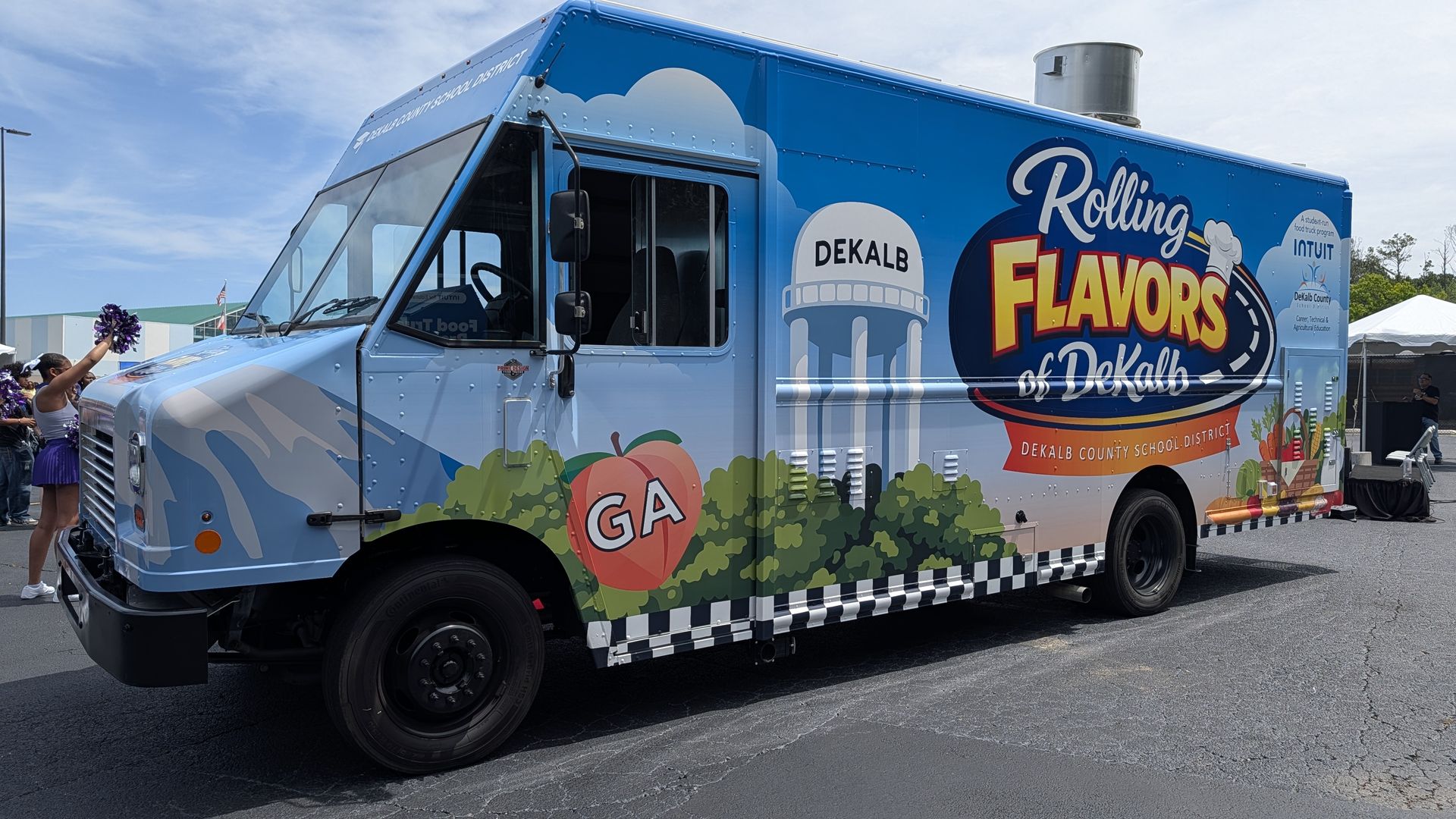 Bright blue food truck with the words "Rolling Flavors of Dekalb" and Dekalb County School District logo, featuring fruit graphics; cheerleaders with purple pom-poms stand nearby at an outdoor event.