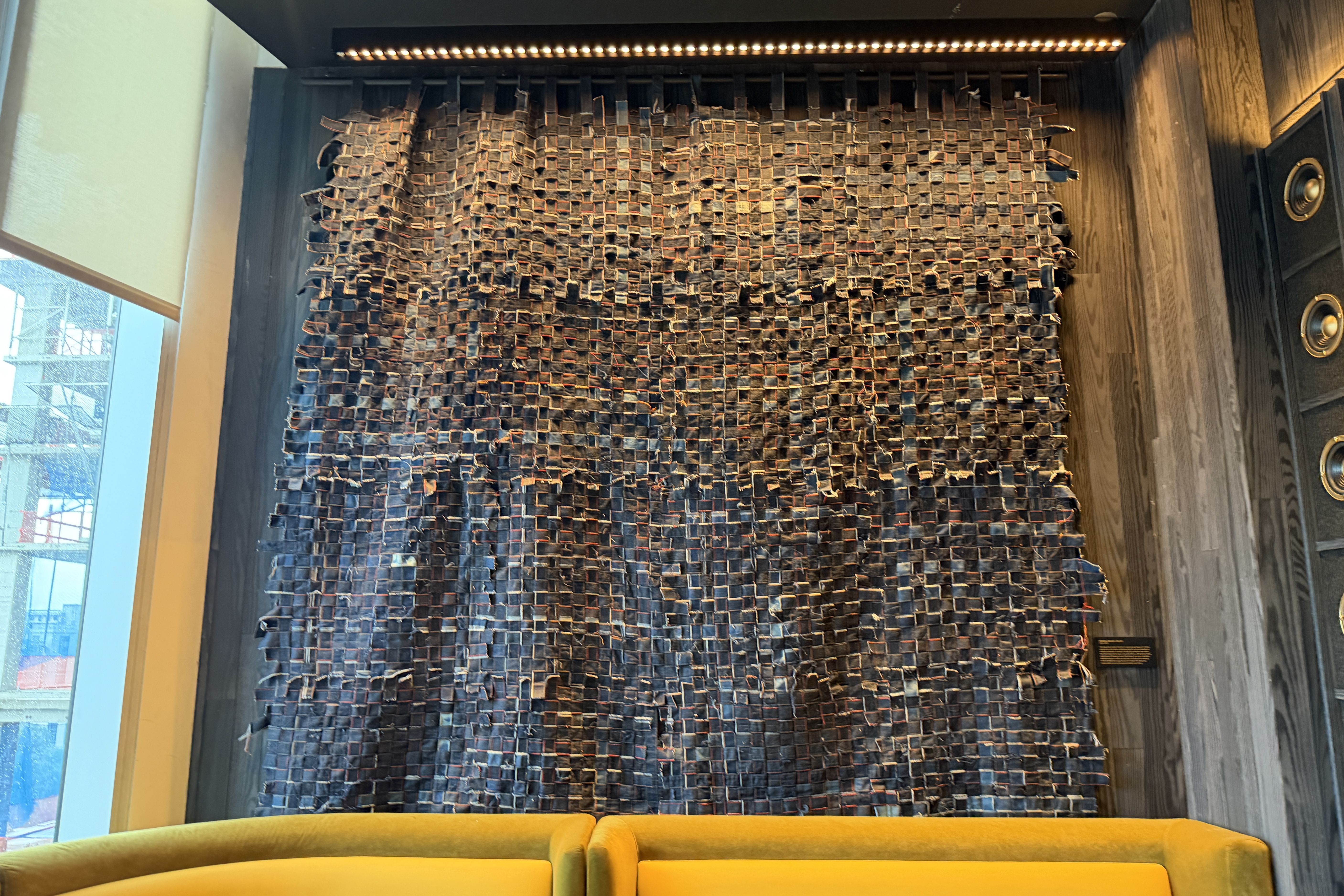 Cozy modern lounge featuring a tall dark woven wall installation made of interlaced fabric strips, warm overhead lamp, wood-paneled walls, and a bright yellow sofa at the bottom.