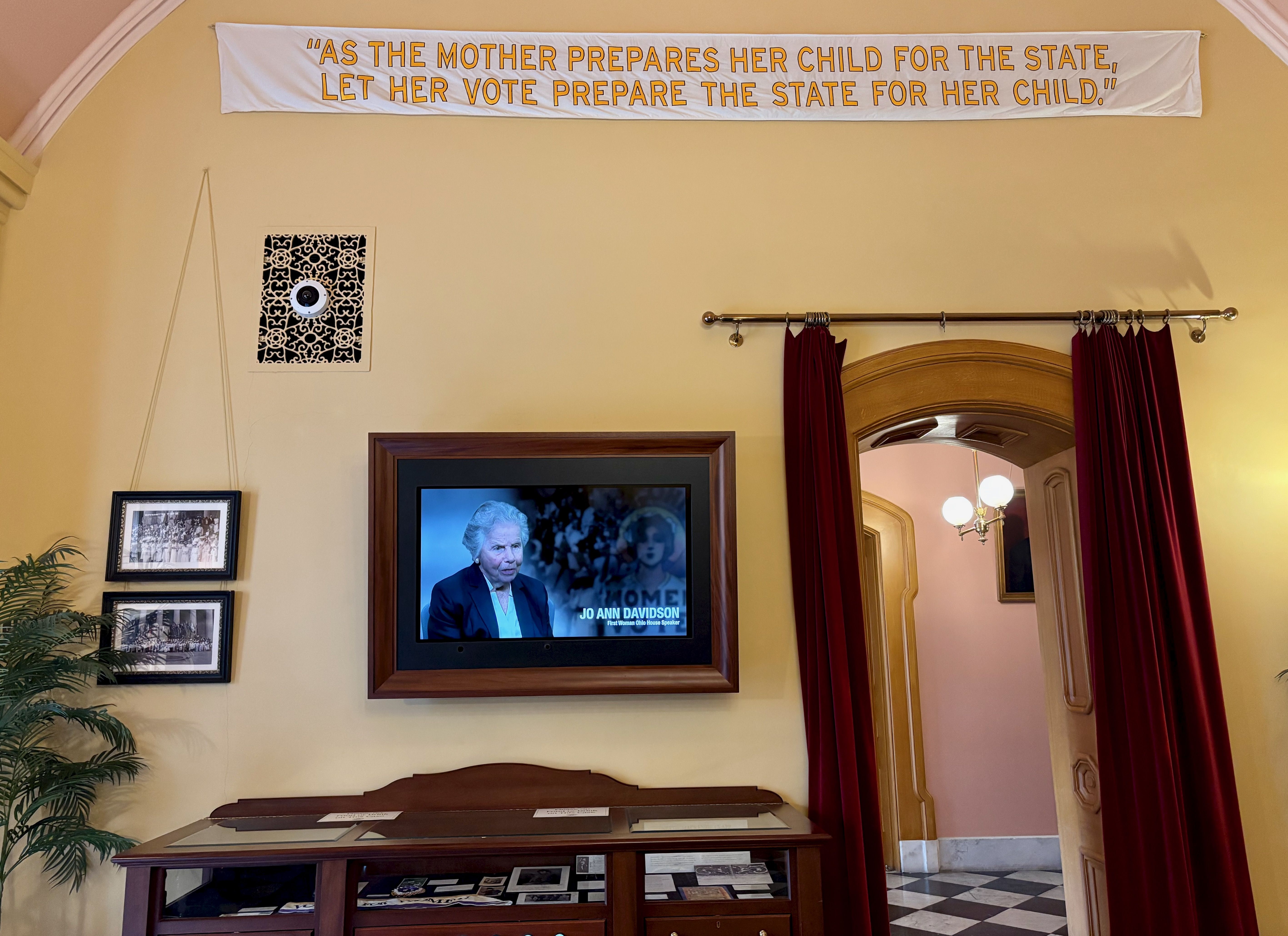 The gallery entrance featuring a TV and a banner that reads "as the mother prepares her child for the state, let her vote prepare the state for her child"