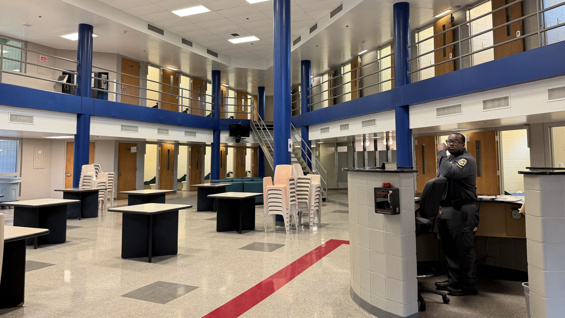 Interior of a two-level lobby with bright white ceiling, blue pillars, and metal railings. Ground floor features stacked chairs and tables; a security guard sits at a curved reception desk on the right.