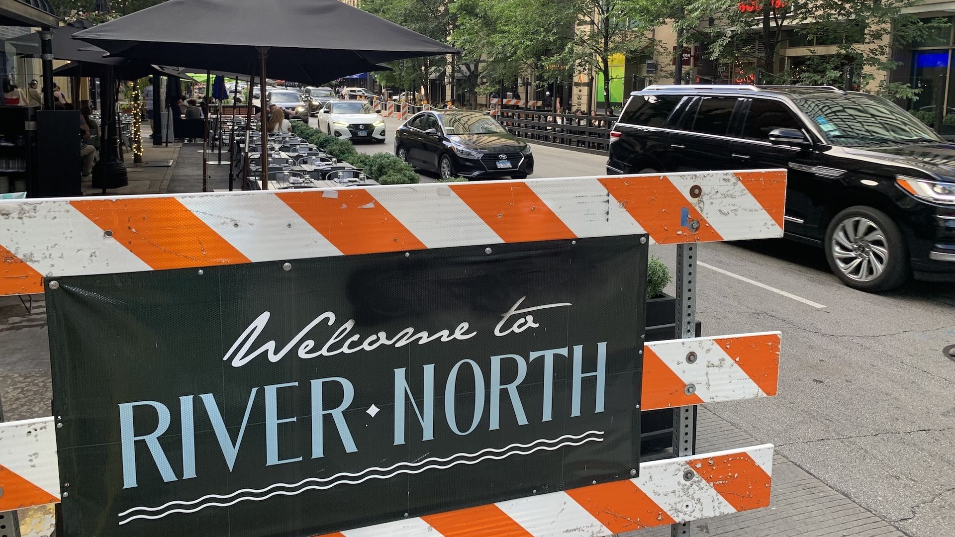 Orange and white traffic sign with "Welcome to River North" black banner on it in front of street with cars and table umbrellas.