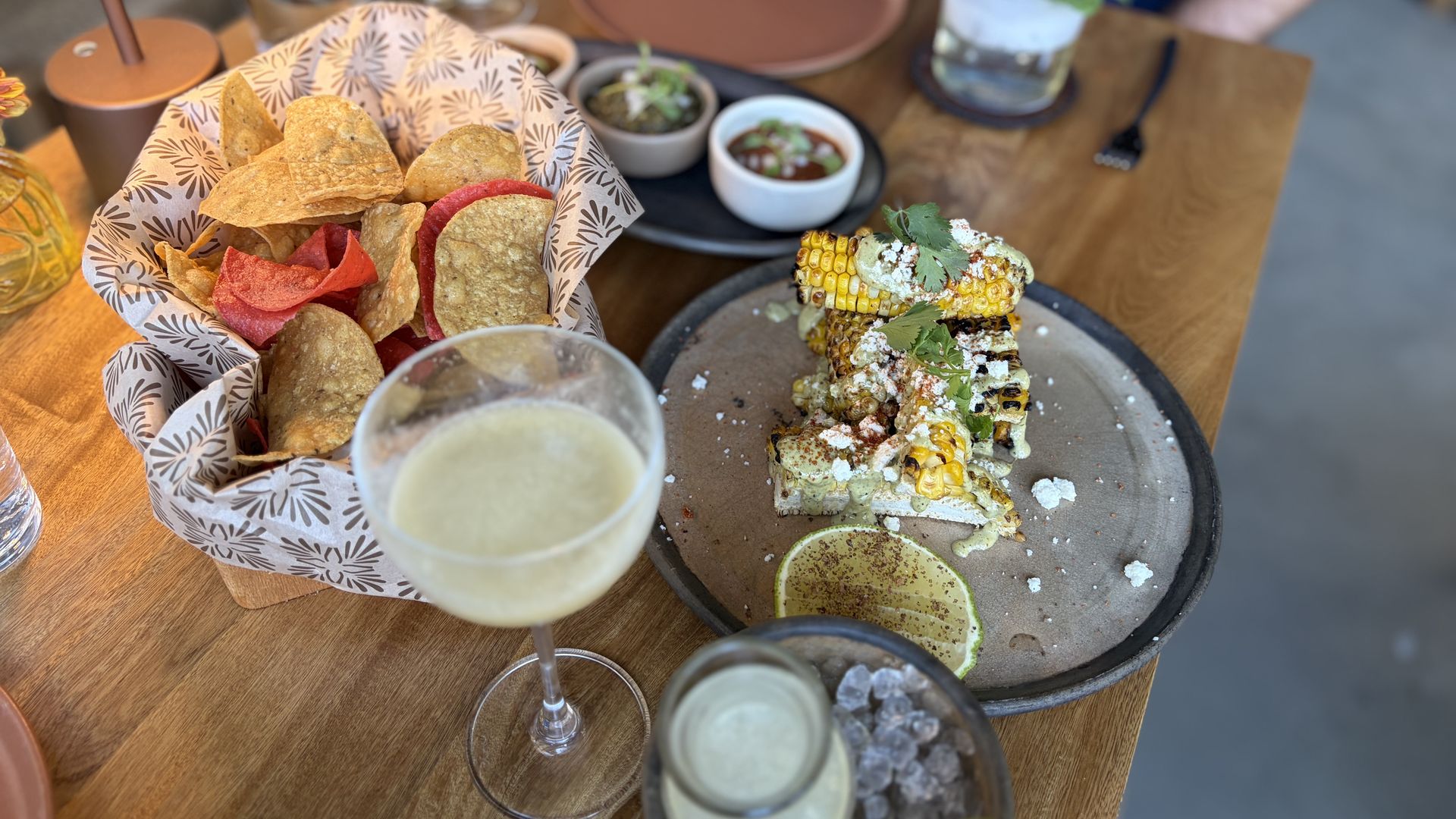 Basket of tortilla chips in patterned paper, two small bowls of dip, and a plate of grilled corn topped with herbs and cheese. Limes and drinks on a wooden table with a blurred figure in blue in the background.
