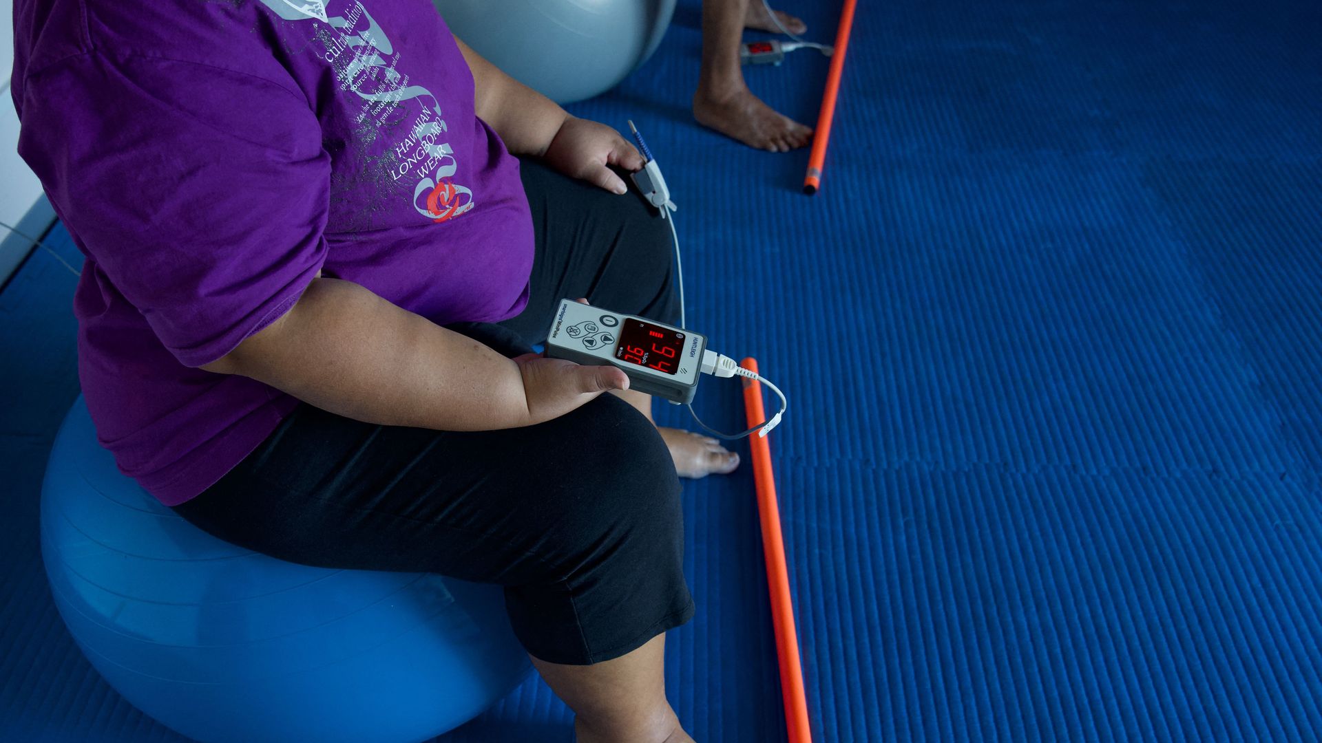 An overweight woman in a purple t-shirt checks her vitals with a small digital machine. 