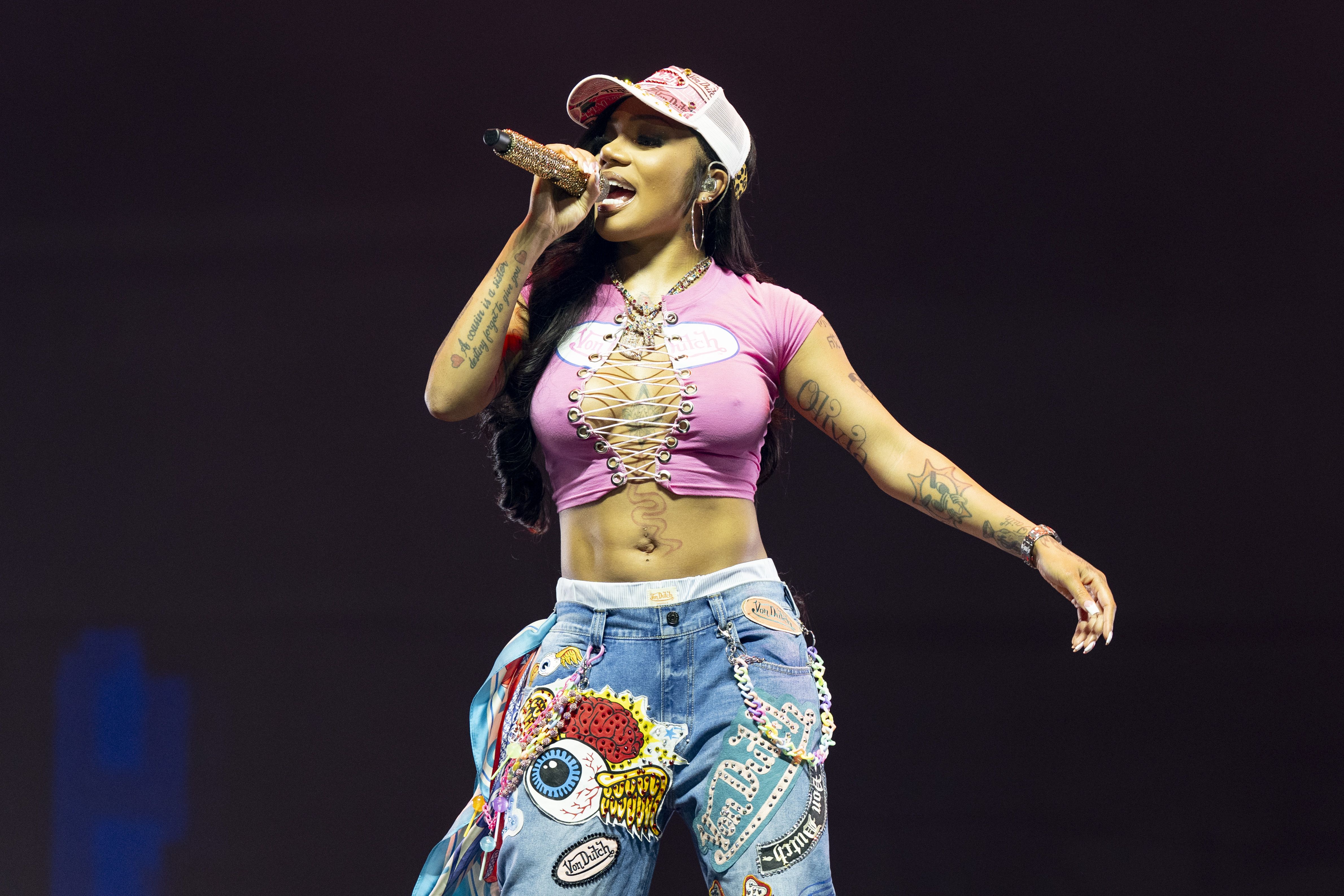 Female performer singing on stage wearing a pink lace-up crop top, decorated blue jeans with colorful patches, a white and pink cap, and holding a glittery microphone against a dark background.
