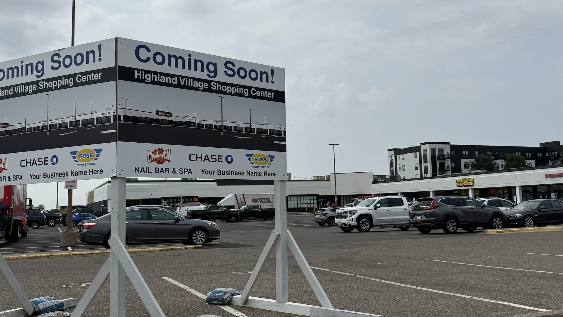 Billboard reads "Coming Soon!" for Highland Village Shopping Center; shows row of storefronts, a parking lot full of cars, and a distant modern building under a cloudy sky.
