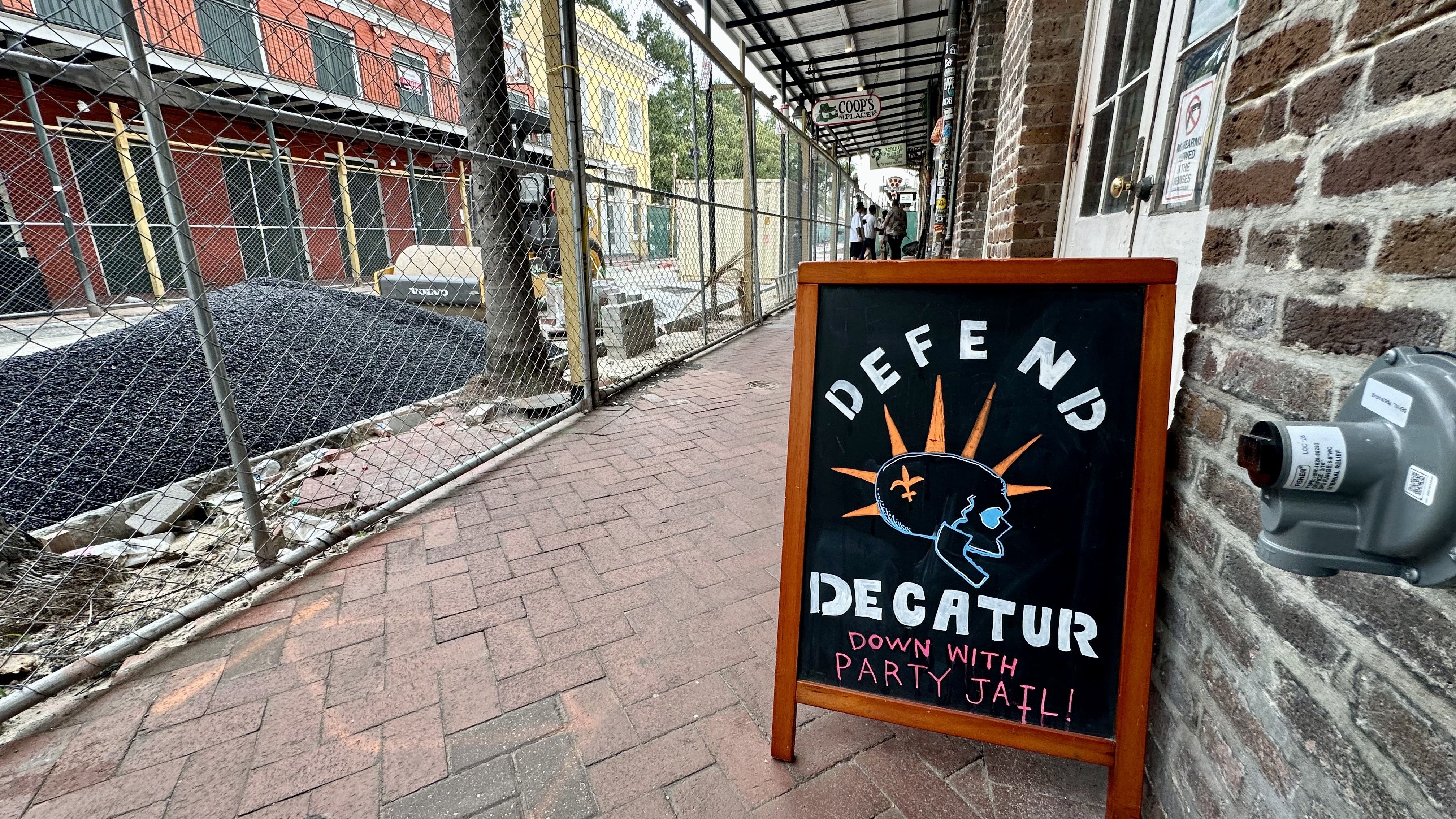 City sidewalk with a chain-link fence on the left and brick buildings behind. A wooden-framed chalkboard sign on the right shows an orange sunburst and the words DOWN WITH PARTY JAIL!