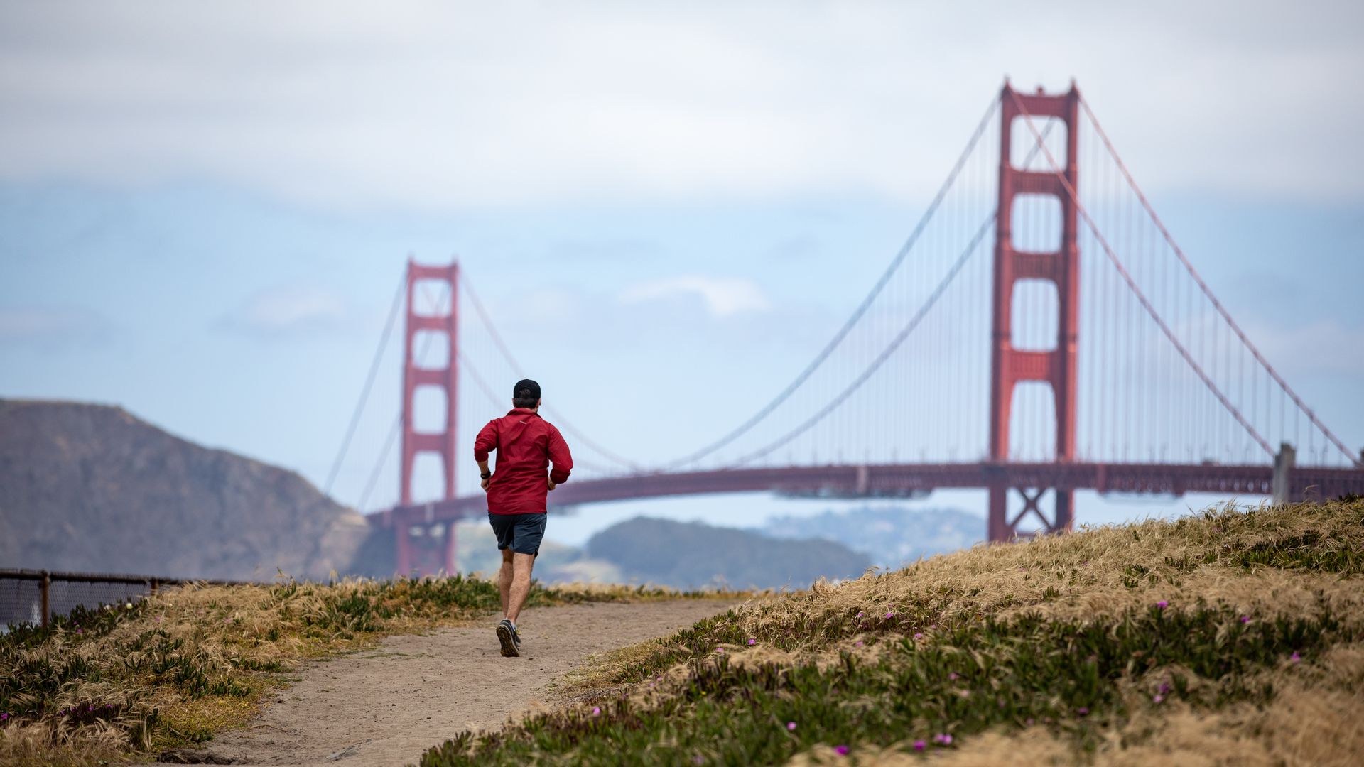 A jogger in a red jacket runs along a dirt path with the Golden Gate Bridge visible in the distance under a cloudy blue sky.