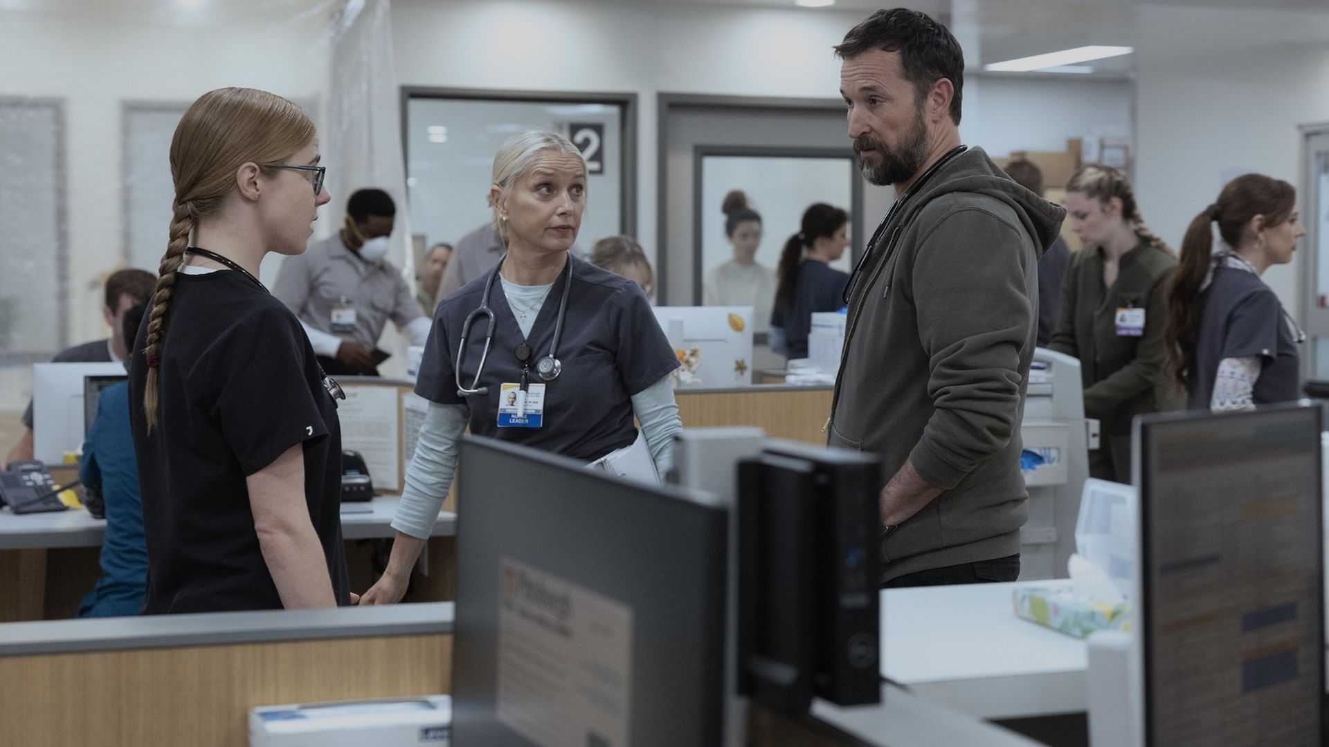 Hospital reception: a bearded man in a green hoodie speaks with a senior nurse wearing a stethoscope, while a young nurse with glasses listens; desks, computers, and staff in the busy background.