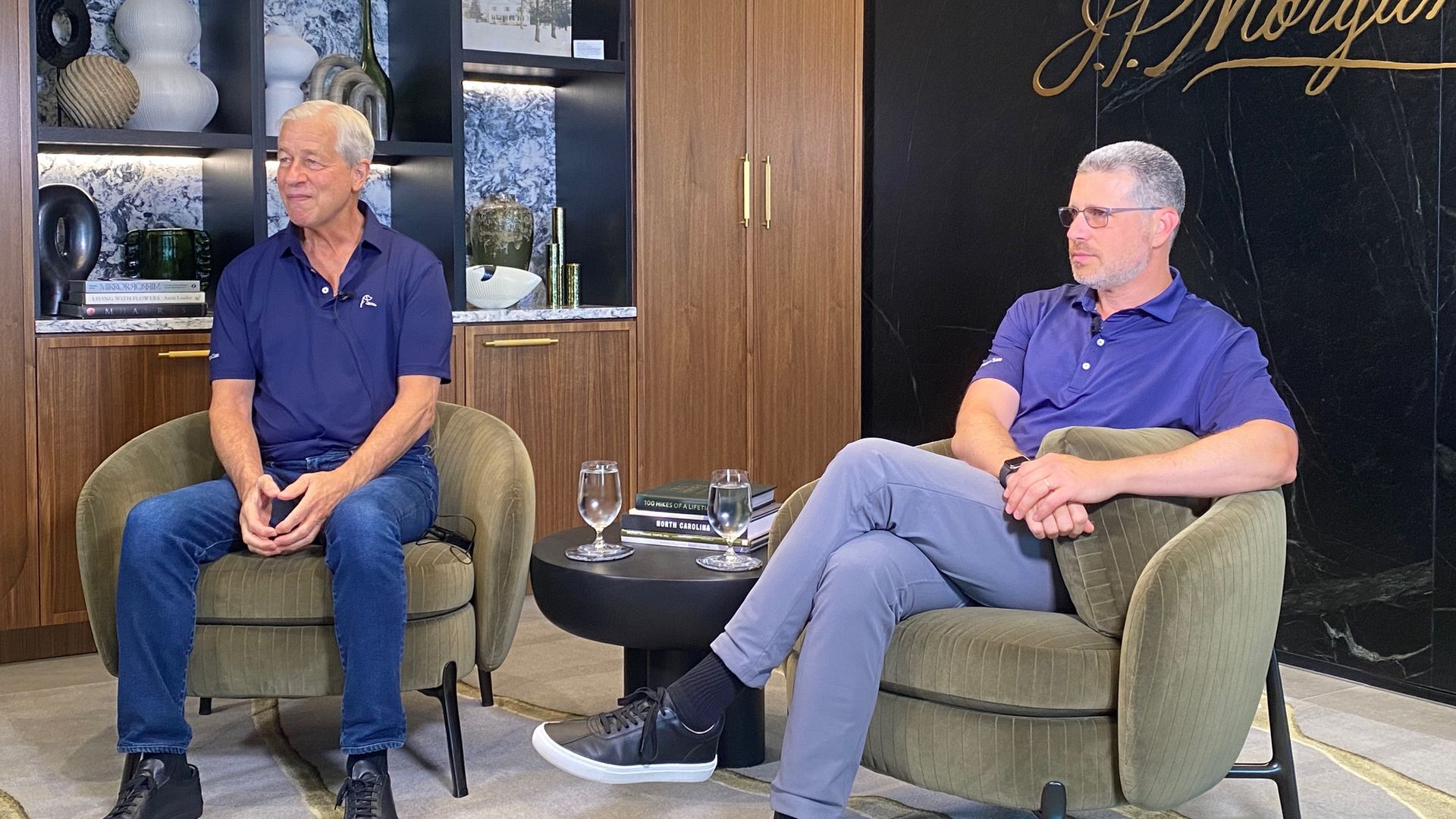 Two men in blue shirts sitting on green armchairs in a room with wooden shelves and a black wall with gold "J.P. Morgan" sign, and a small table with water glasses and books.