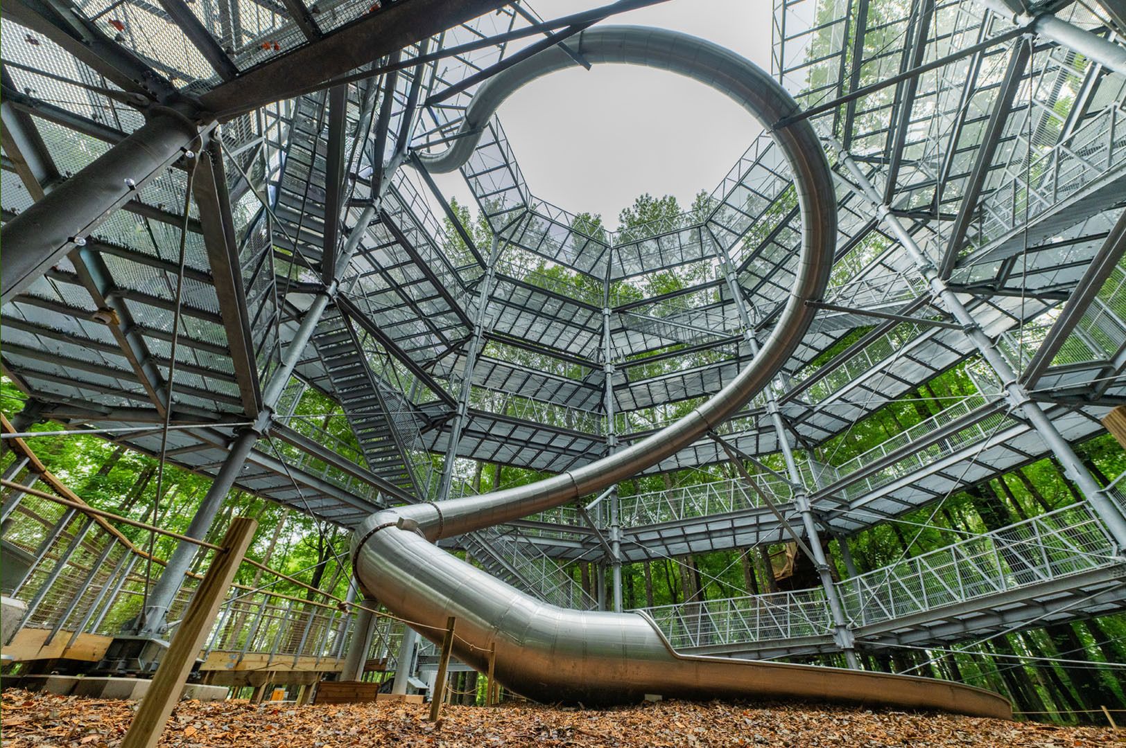 A large, multi-level metal treehouse structure with stairs and walkways surrounds a tall metal slide twisting from the top to the forest floor covered in brown leaves.