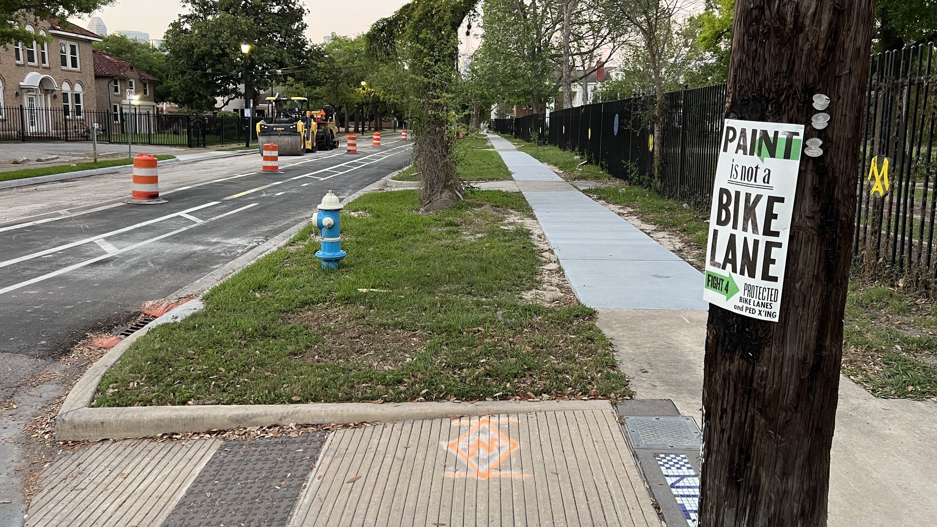 City street under construction with orange cones and distant yellow machinery. A blue hydrant sits on a grassy median; pale blue sidewalk runs beside a black fence near a tree. A sign reading "Paint is not a bike lane" sits on a utility pole