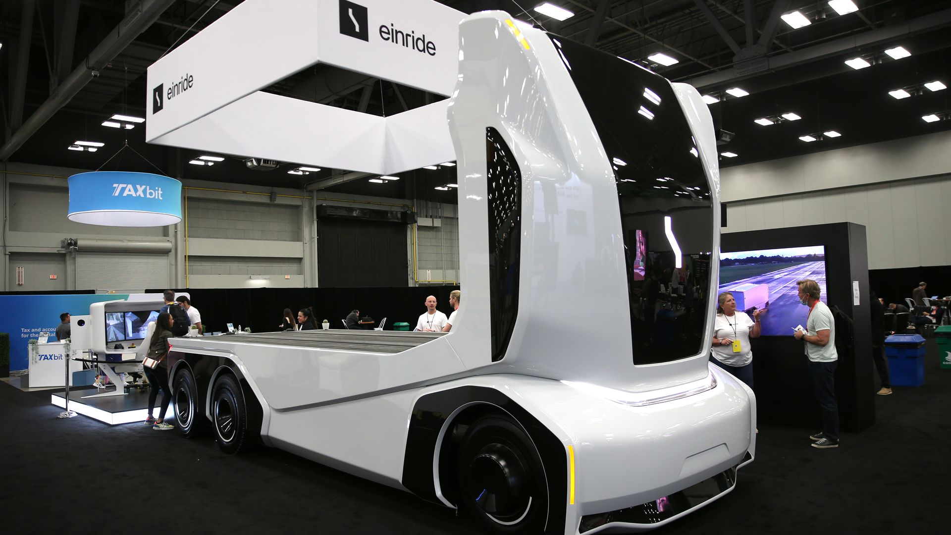 White futuristic electric truck with smooth rounded edges and large black windshield on display at an indoor tech expo, with people and exhibitor booths in the background.