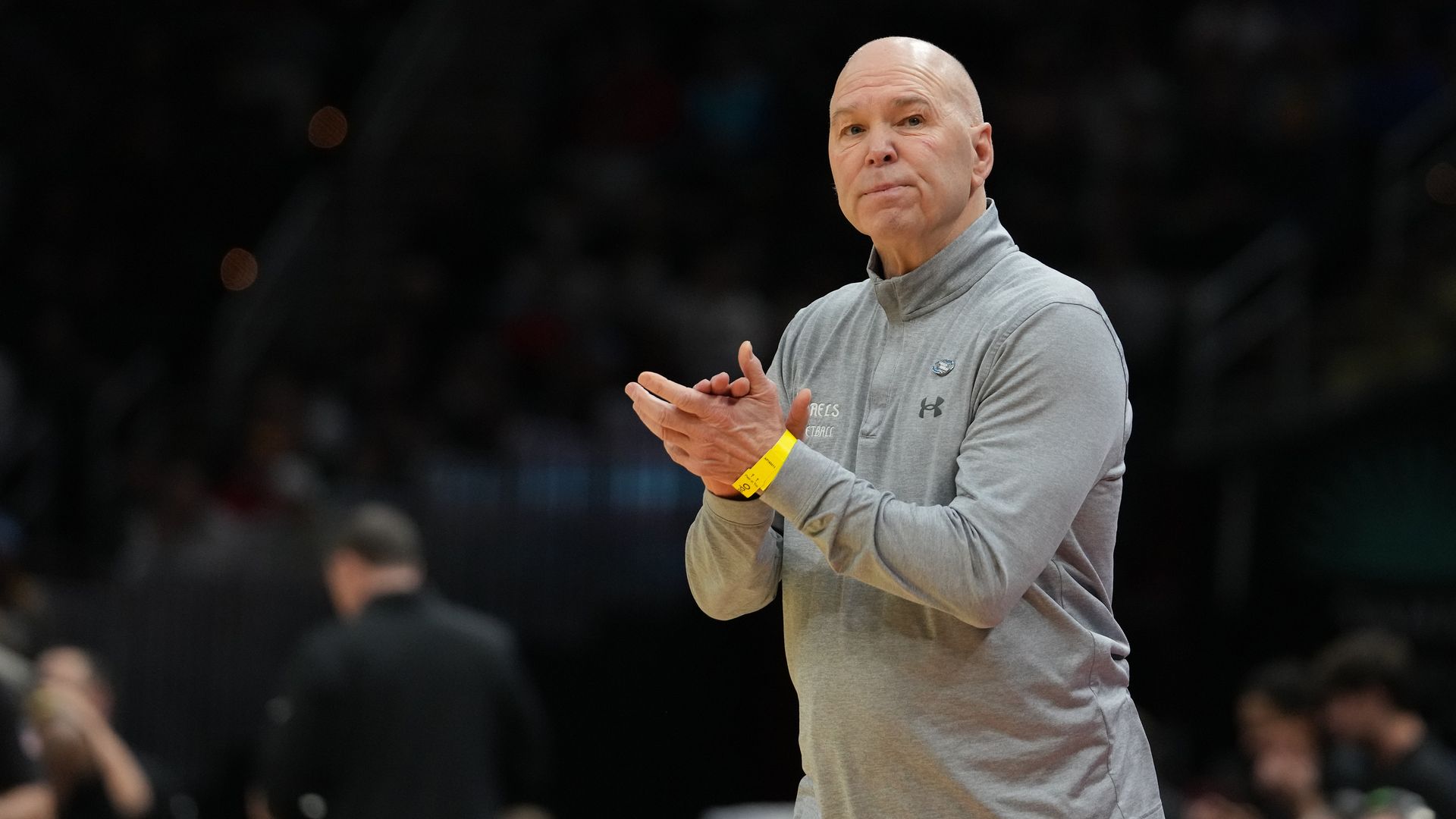 A bald man wearing a light gray athletic pullover claps at a basketball game, with a yellow wristband on his right wrist, while a blurred crowd watches in the background.