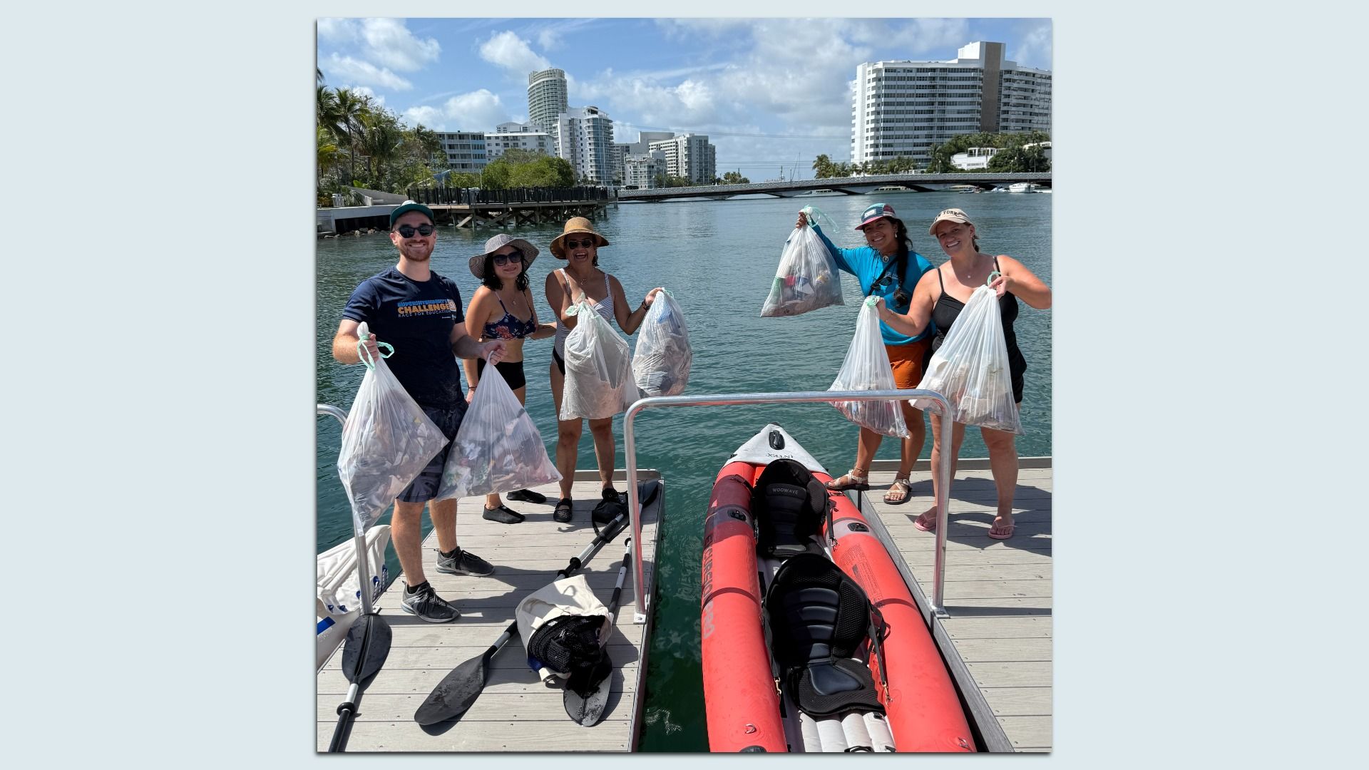 Five people on a dock holding clear bags filled with trash, standing beside a red inflatable kayak on a sunny day with buildings and water in the background.