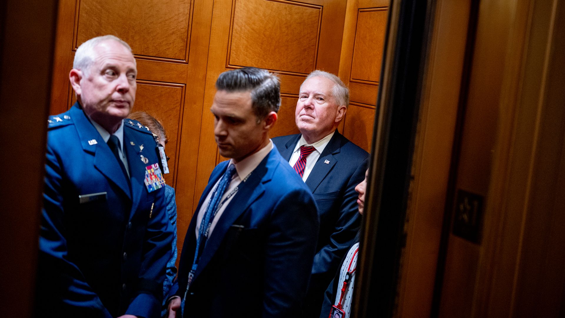 Five people crowd into an elevator, decorated with warm-toned wood. Three of the men visible are in blue clothing, including one military uniform.