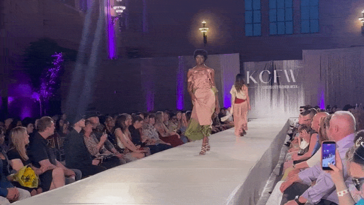 Runway fashion show with two models in peach dresses; one has lime-green underlayer and lace-up sandals. Purple lighting, audience on both sides, some people filming; backdrop with large white letters.