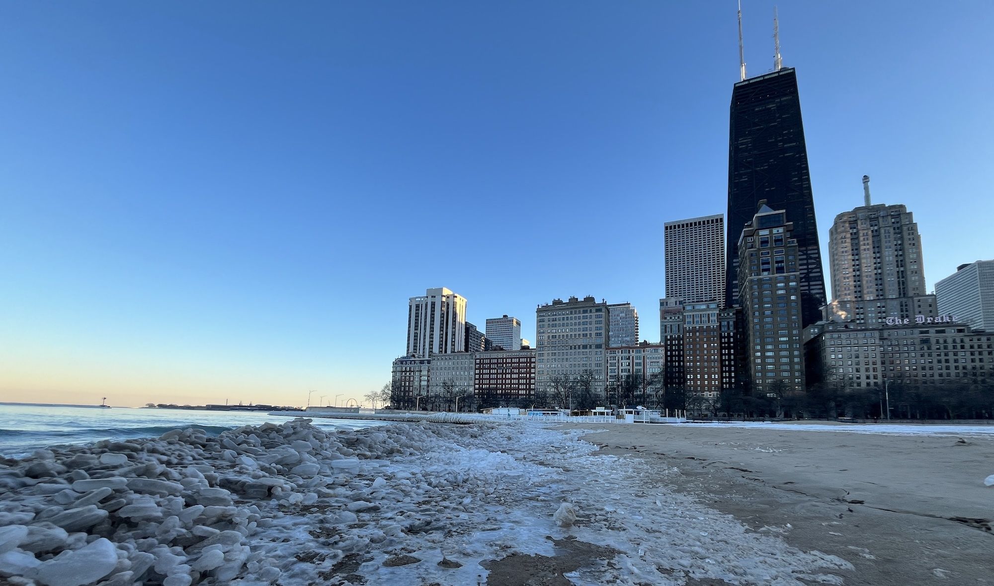 Frozen shoreline with ice chunks and sandy Oak Street beach, city skyline with tall buildings including a black skyscraper and "The Drake" hotel, under clear blue sky at sunrise or sunset.