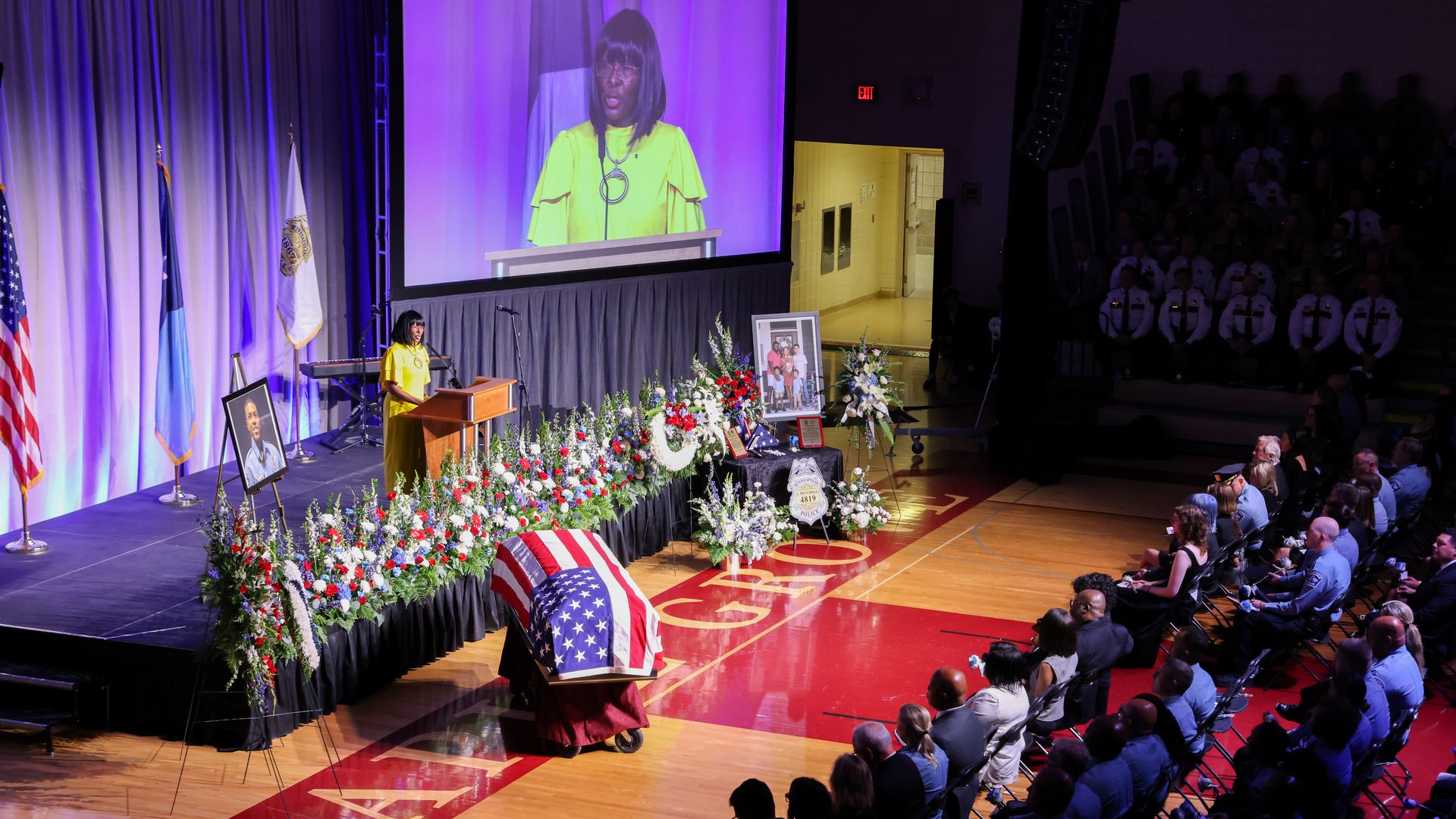 A woman in a yellow dress speaks on a stage with a large video projector in a darkened gymnasium with blue-clad dignitaries seated in rows of folding chairs on the floor. A flag-draped casket sits in front of the speaker.