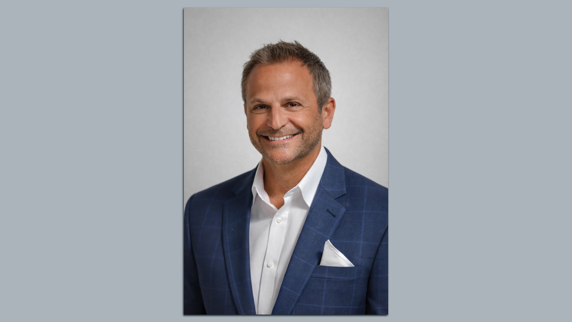 Smiling middle-aged man with short gray hair and beard wearing a blue checkered blazer, white shirt, and white pocket square against a plain gray background.