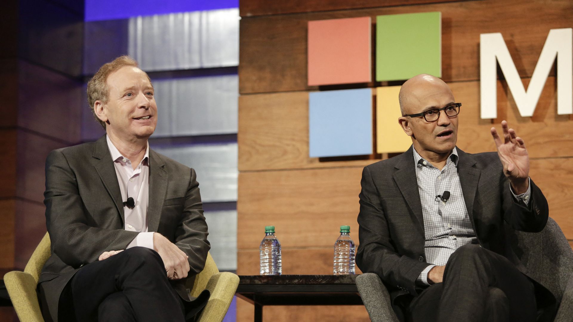 Microsoft CEO Satya Nadella and president Brad Smith sit on a stage in front of the Microsoft logo.