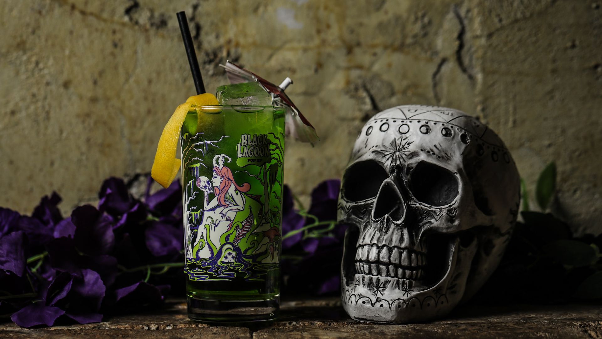 Green cocktail in a Black Lagoon glass with lemon peel, umbrella, and black straw, sitting next to a decorated white skull on a rustic surface with purple flowers and a textured wall background.