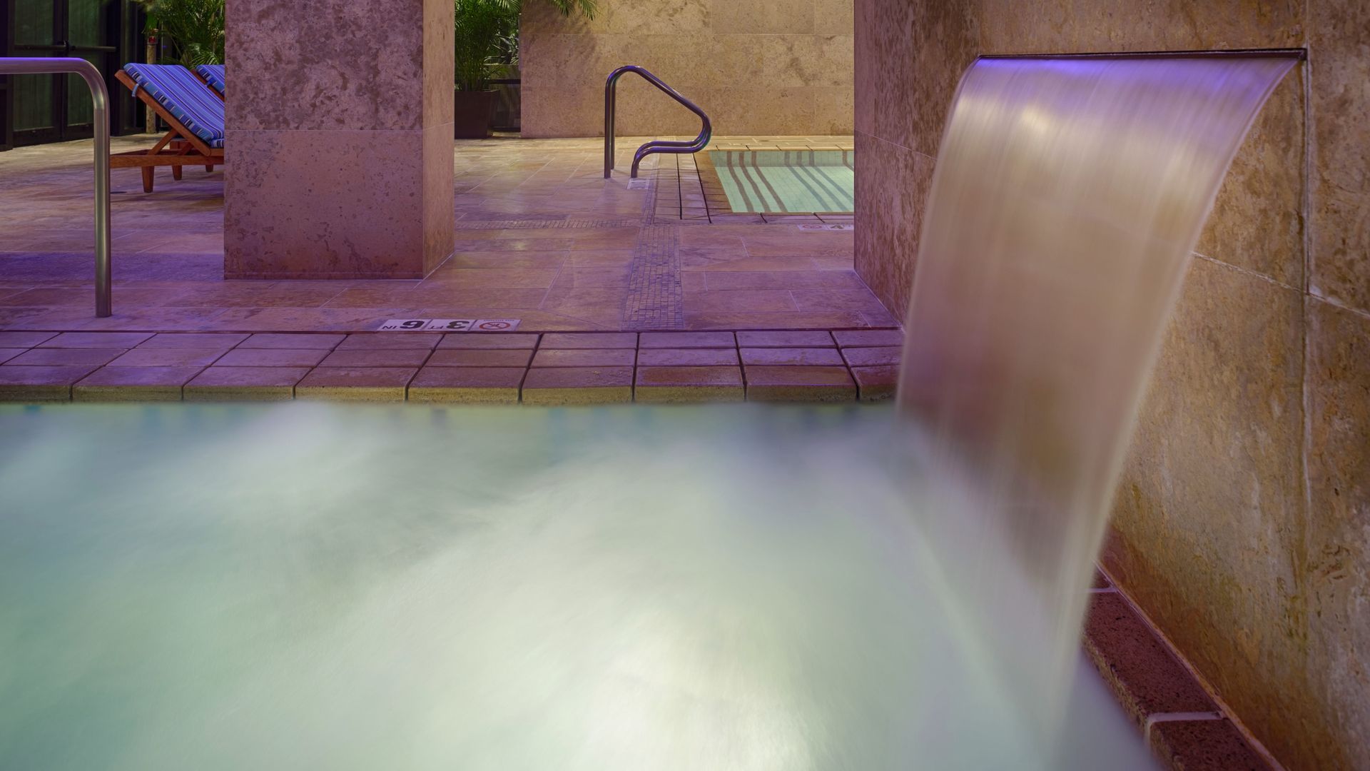 Indoor pool area with a stone waterfall feature pouring water into a misty pool, beige tiled floors and walls, metal railings, and blue lounge chairs in the background.