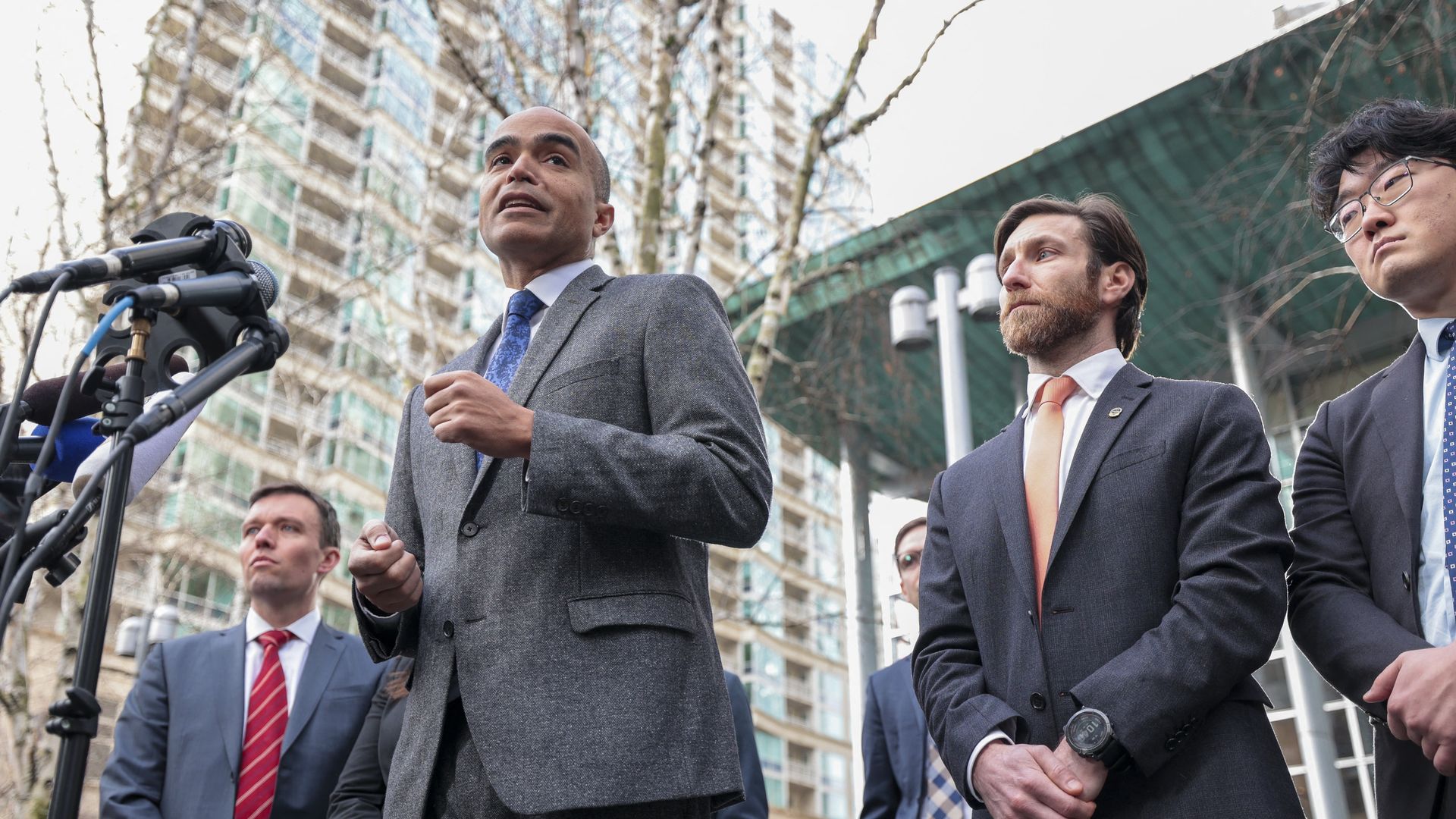 Nick Brown speaks at a podium with several microphones in front of a downtown building and courthouse, wearing a gray suit. He is joined by three other men in suits.