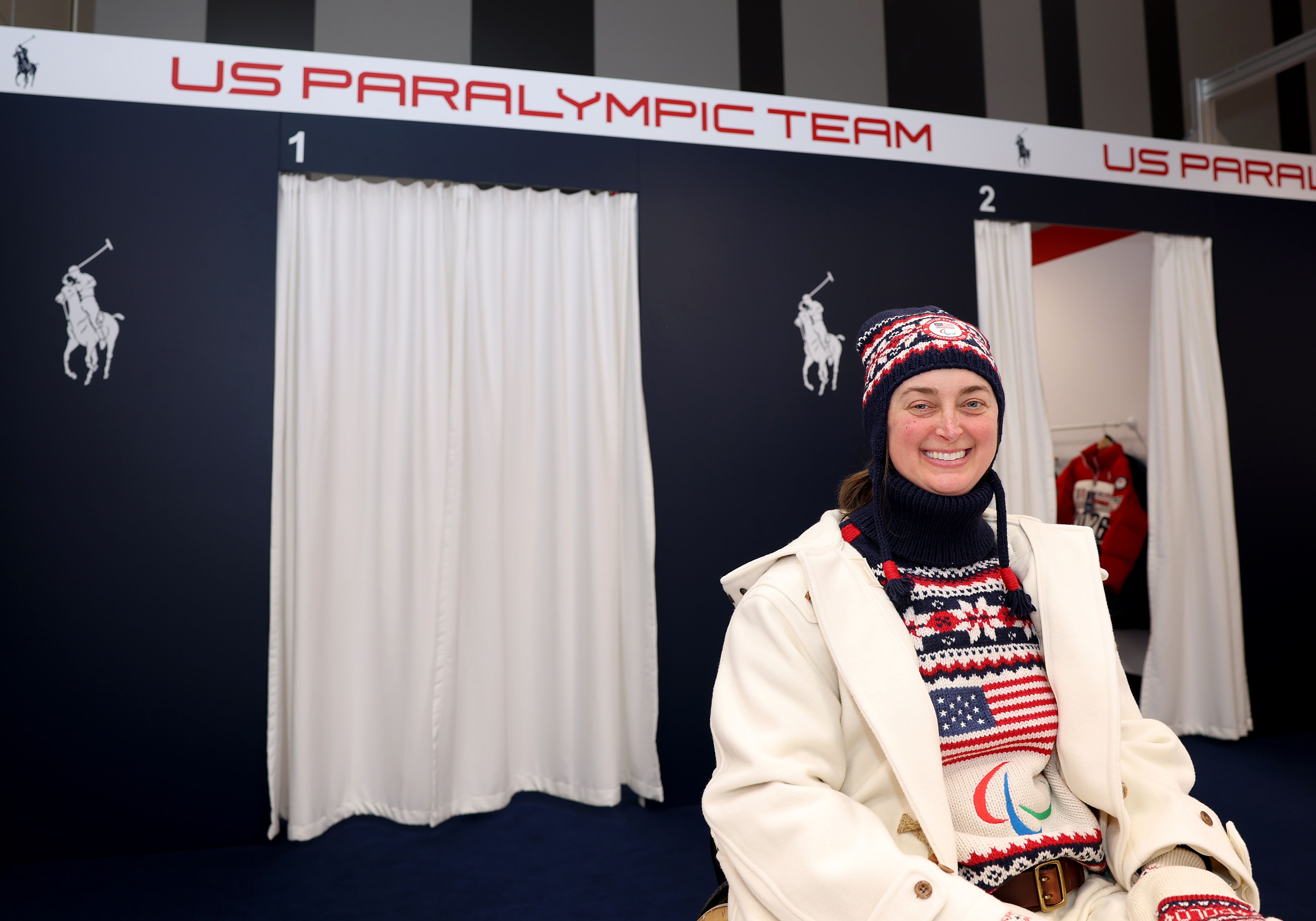 Erin Martin sits in a knit hat, white jacket and USA flag sweater in front of a sign that says "US Paralympic Team." 