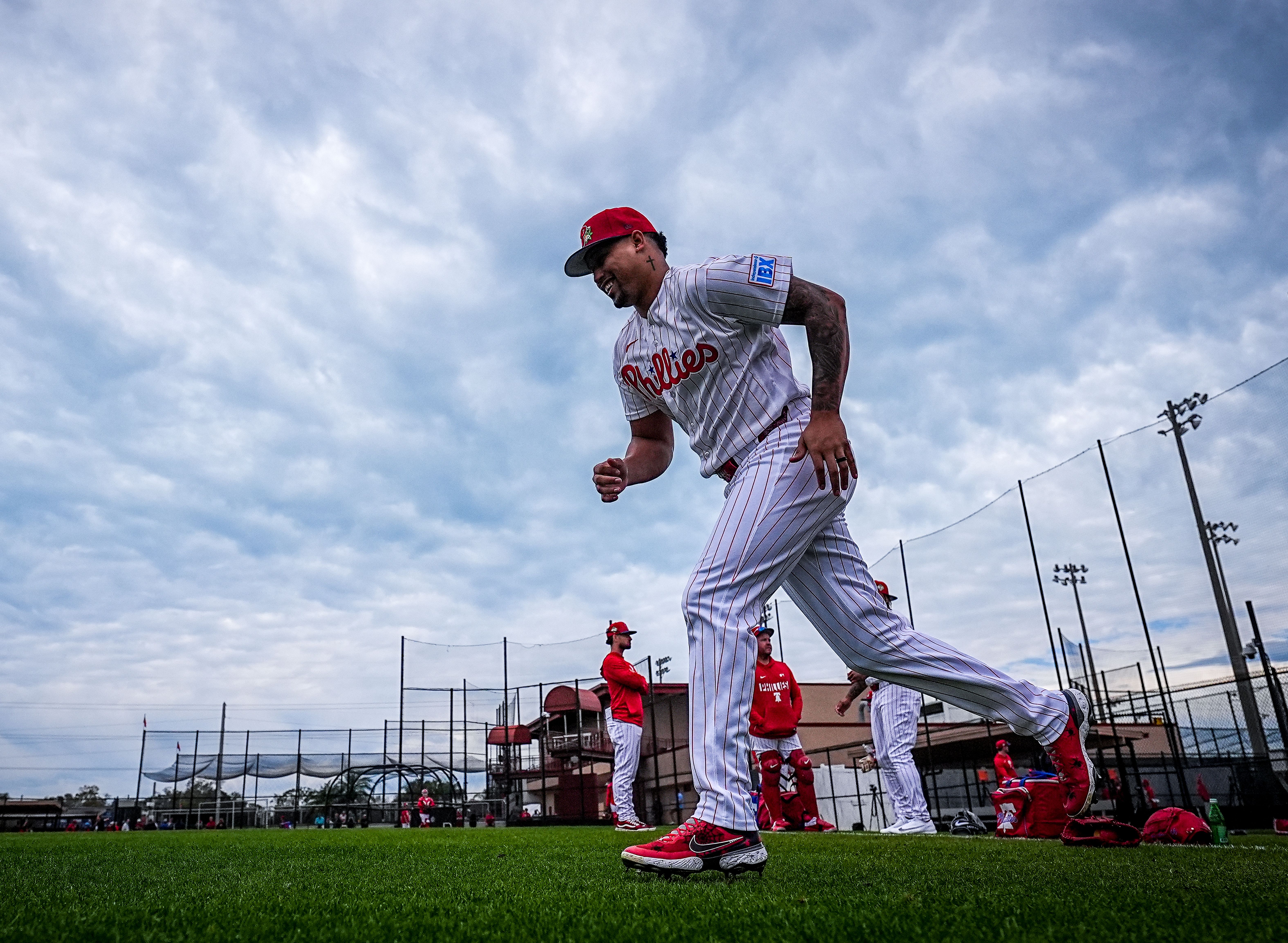A Phillies baseball player in white pinstripe uniform and red cap runs on a green field under a cloudy sky, with teammates and batting cages in the background.
