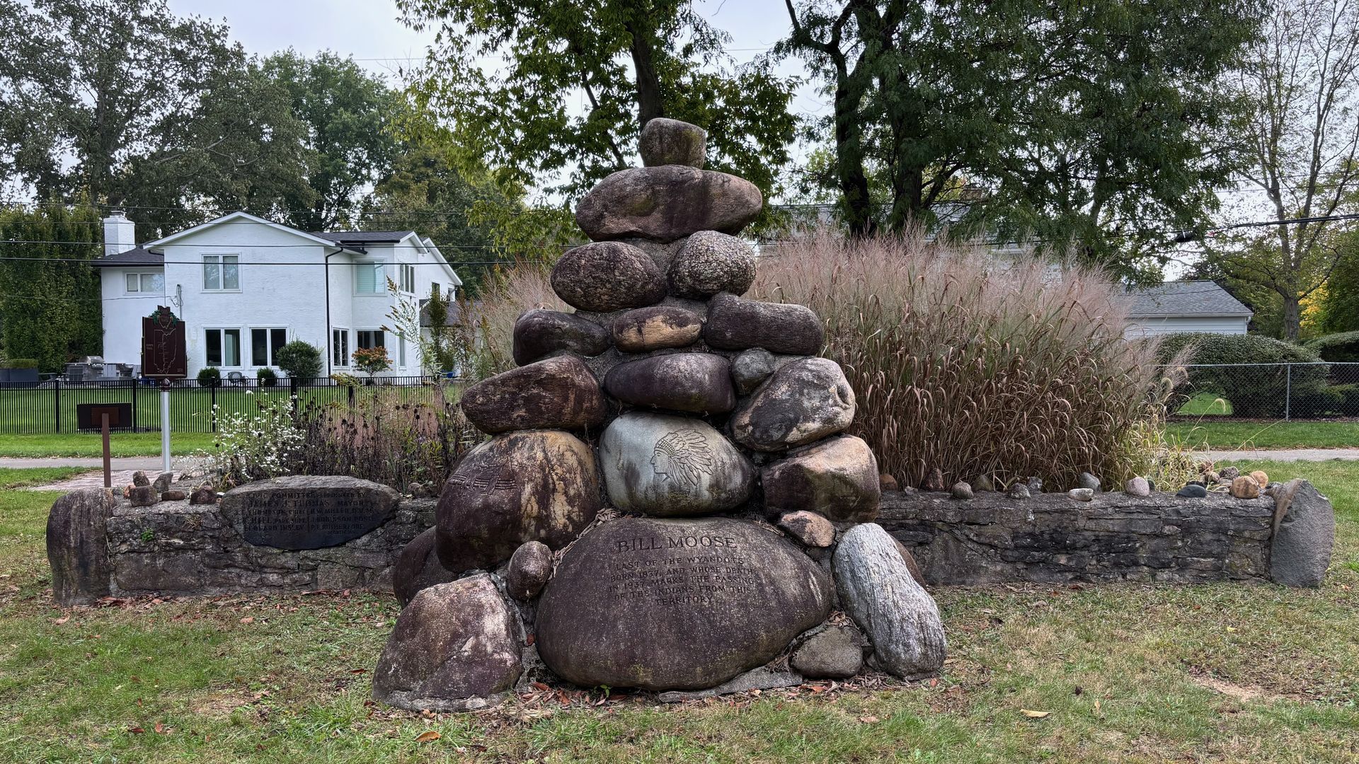 Stone monument with engraved depiction of a Native American chief and inscription about Bill Moose, set in grassy park with trees and white house in background.