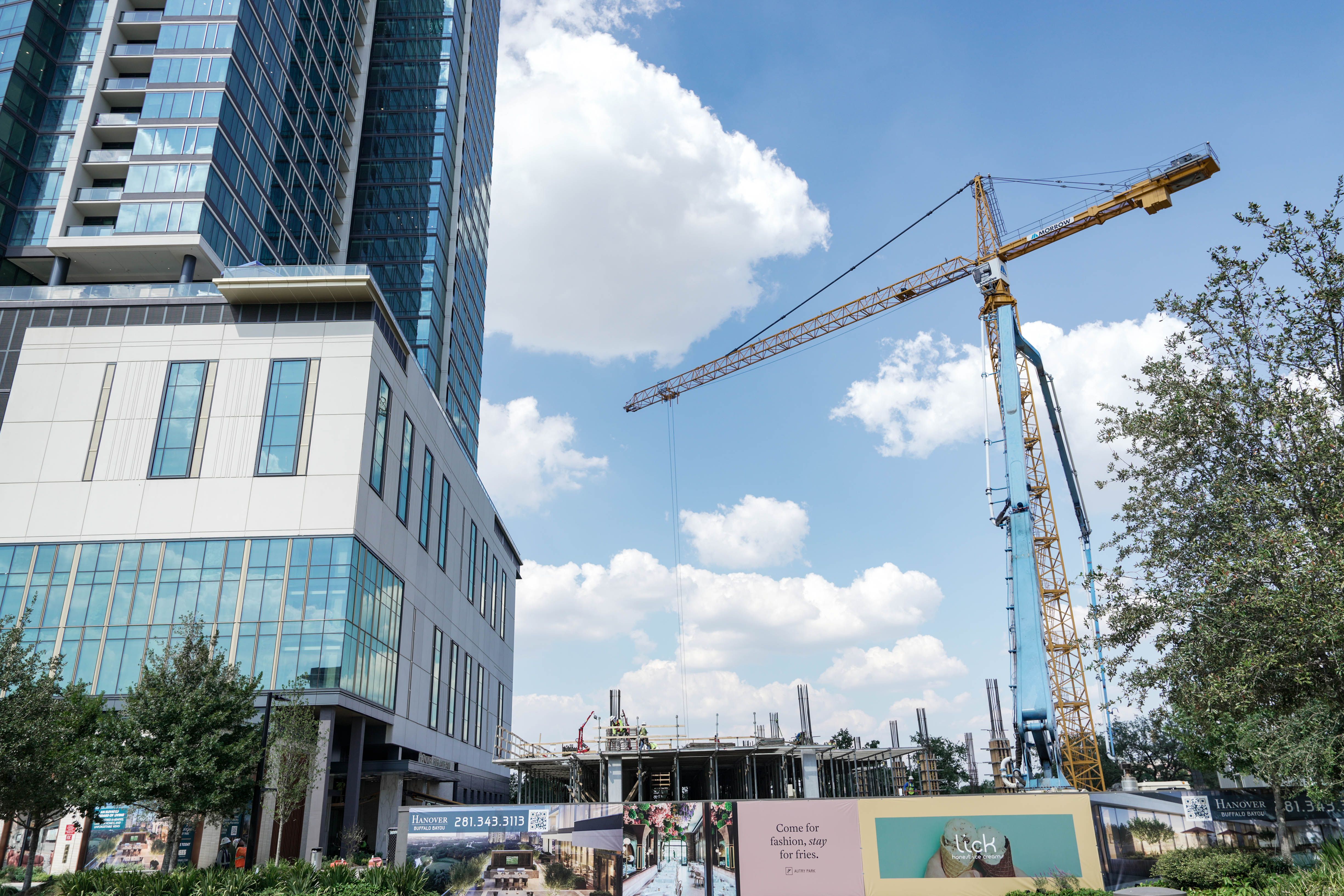 Tall building on the left with glass windows next to a construction site featuring a yellow crane under a partly cloudy blue sky. Some trees and advertising banners are visible.