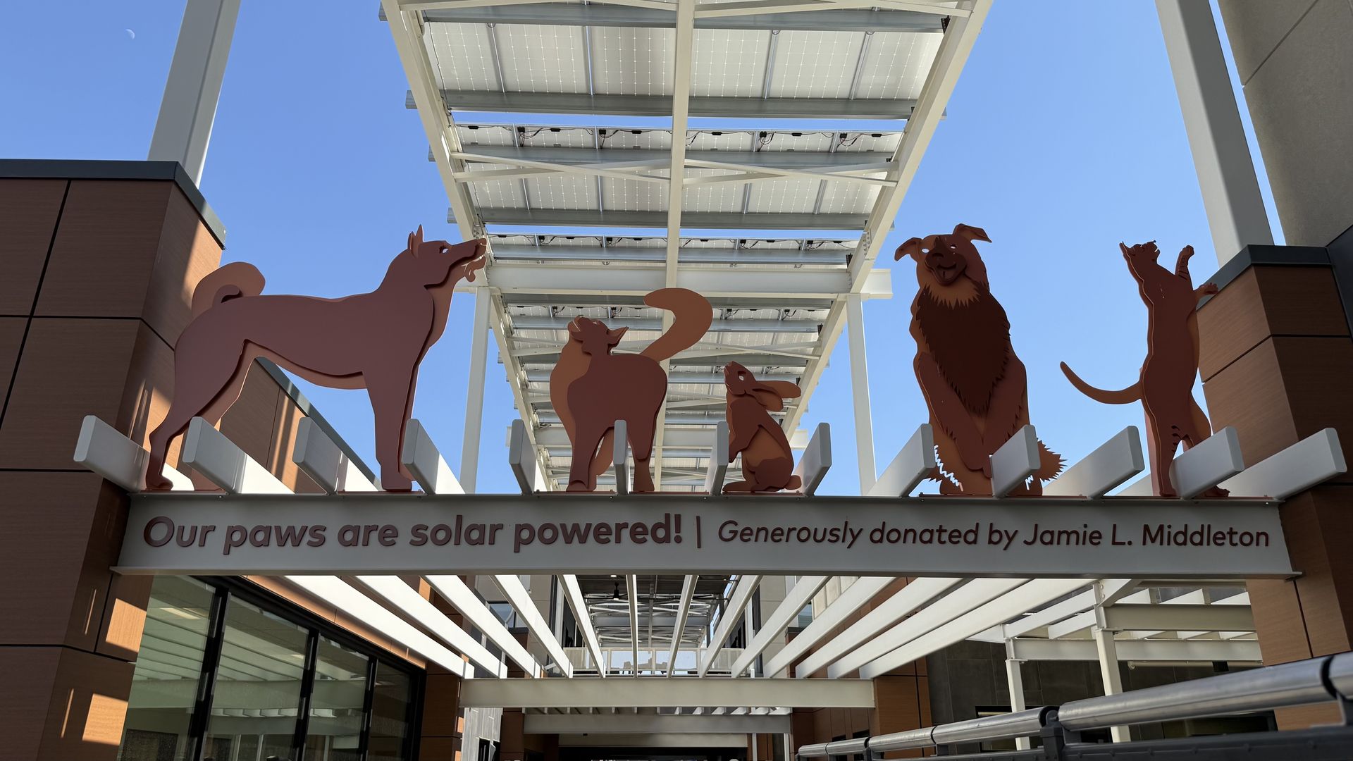 Orange dog, cat and rabbit silhouettes atop a sign that reads, "Our paws are solar powered! Donated by Jamie L. Middleton." The sign is over a walkway at the entrance of a building complex, under a solar panel canopy, with a blue sky above.
