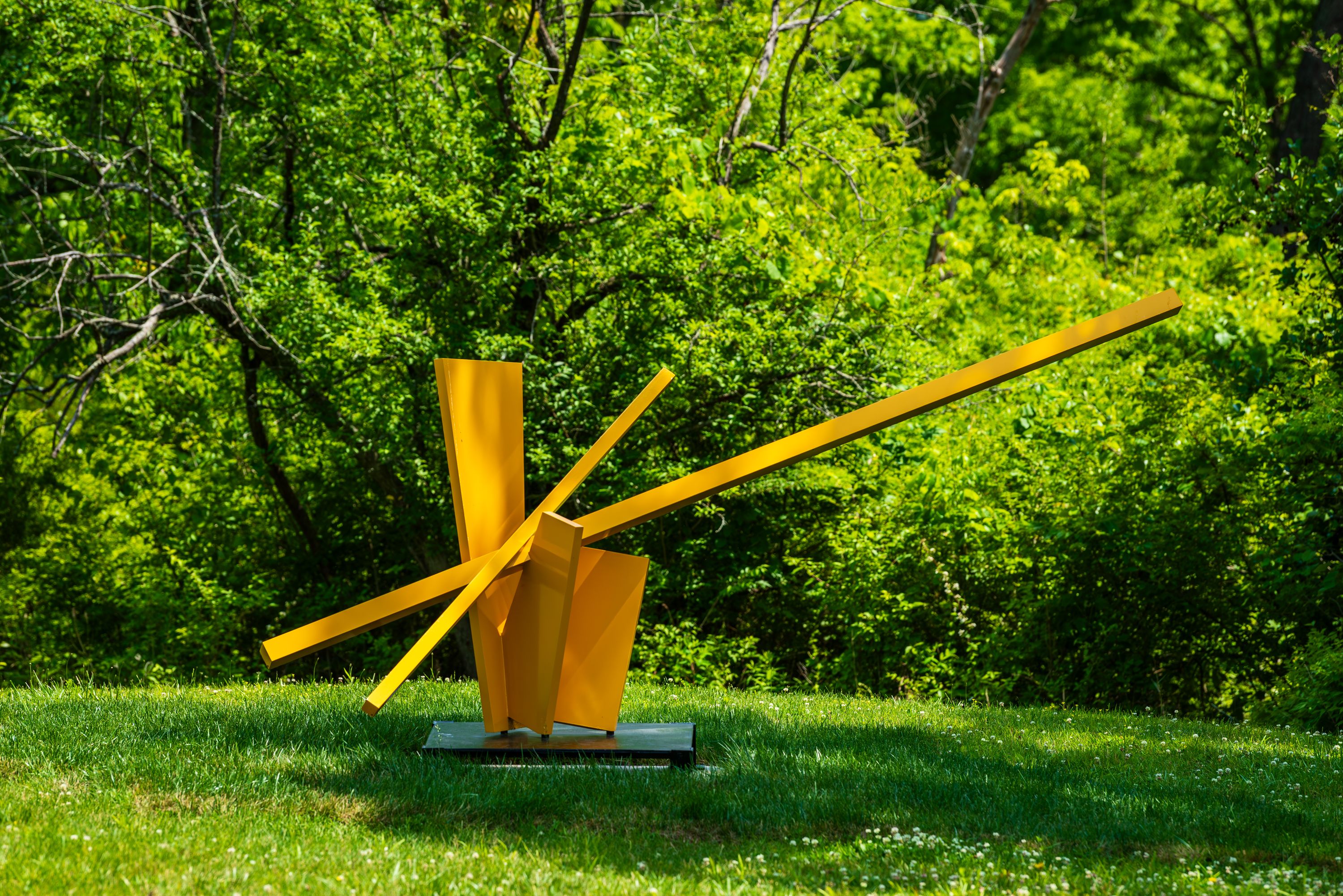 Bright yellow abstract sculpture with intersecting angular beams rising from a square base on a green lawn, set against lush trees in a sunny park.
