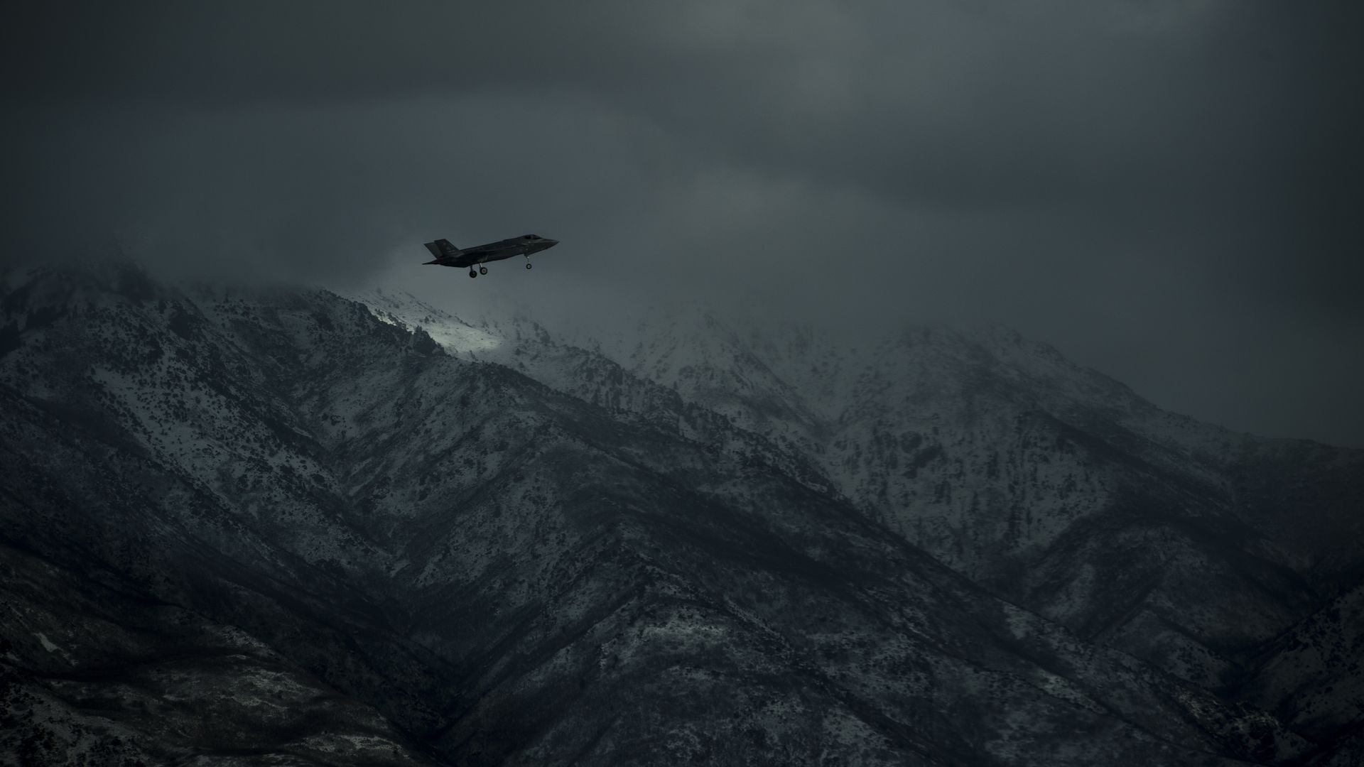 An F-35 fighter flys through darkened skies. A snowy mountain range is seen in the background.
