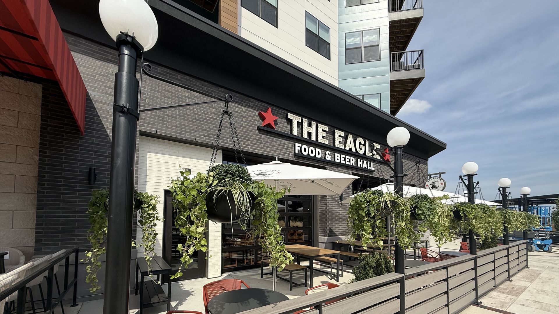 Outdoor seating at The Eagle Food & Beer Hall: gray brick facade, white sign with red stars, hanging plants, black awning, and a modern building with balconies in the background.