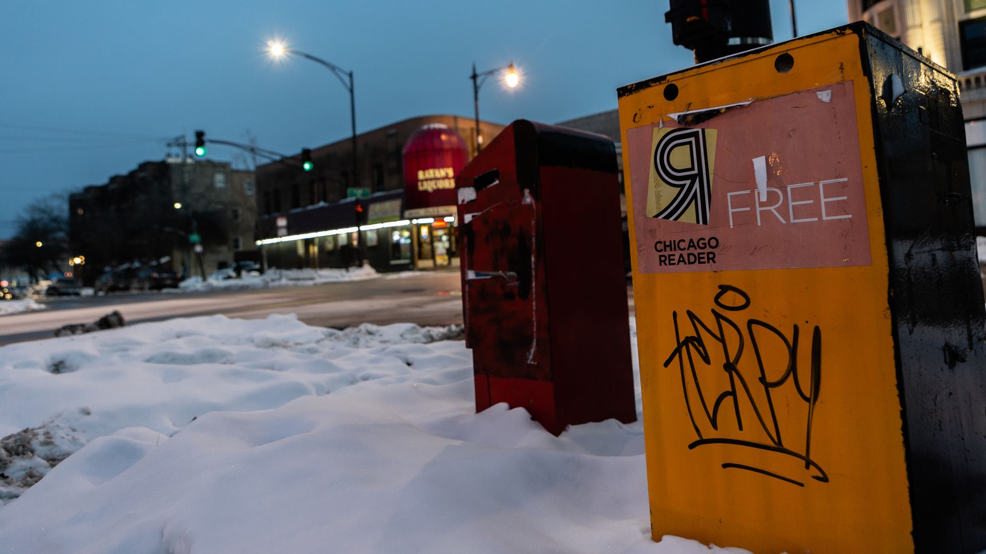 Phot of a newspaper box sitting on the street covered in snow. 