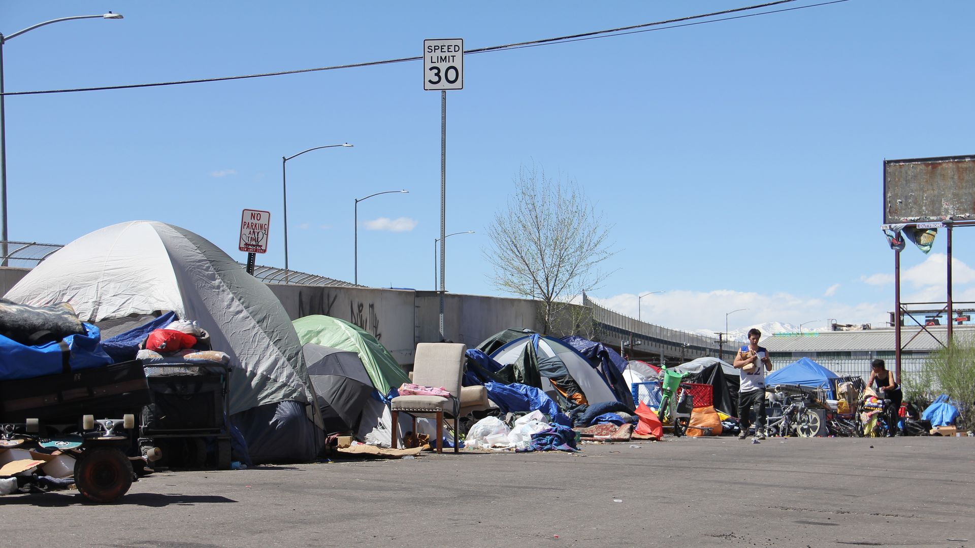Several camping tents of various sizes and colors are lined up against a wall next to a bridge, while one man walks in the foreground, and a woman pushes a cart next to him.