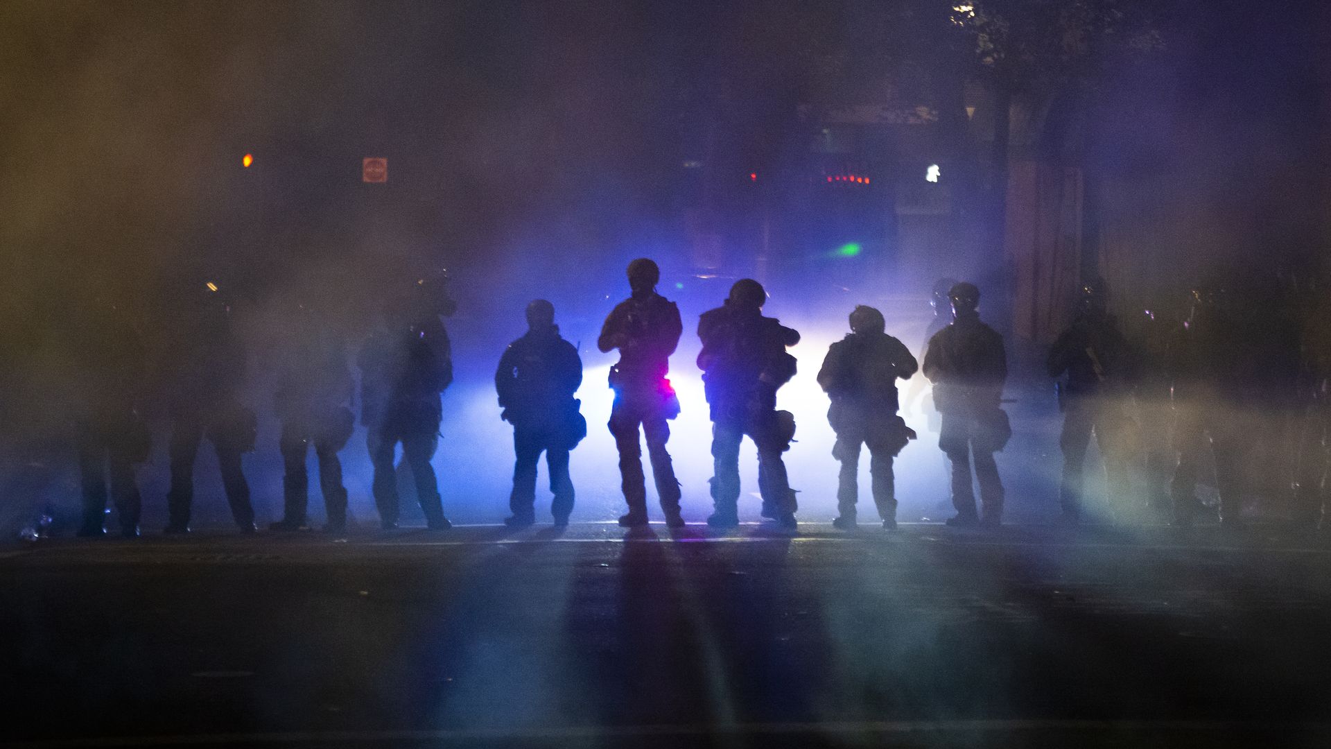 A line of armed officers are backlit by police lights and tear gas
