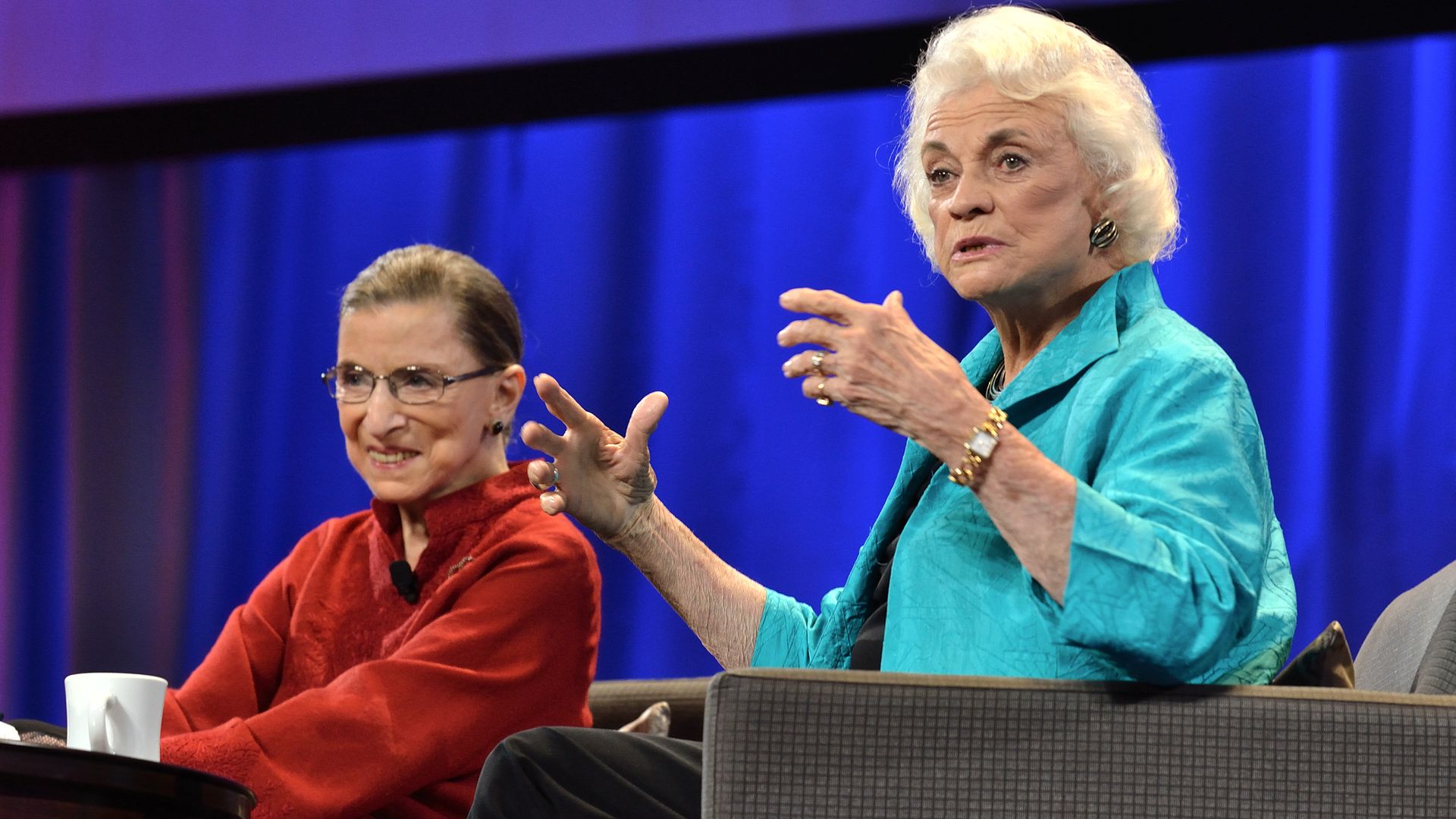 Photo of Ruth Bader Ginsburg in a red button-up shirt and Sandra Day O'Connor in an aqua blazer sitting on stage in a discussion