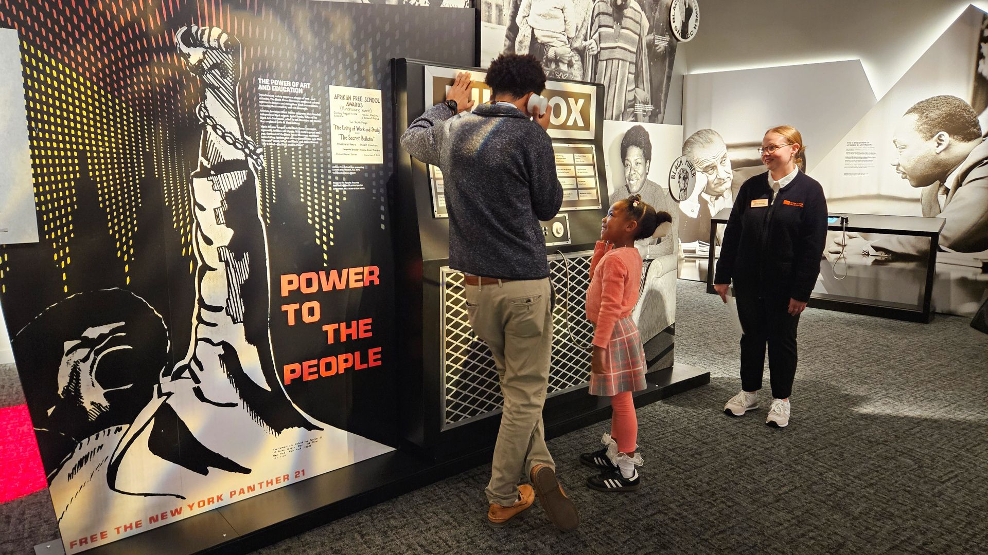 People in a museum exhibit with a large poster showing a raised fist breaking chains and the words "POWER TO THE PEOPLE" in red letters. A man listens to an audio device while a girl and a woman watch.