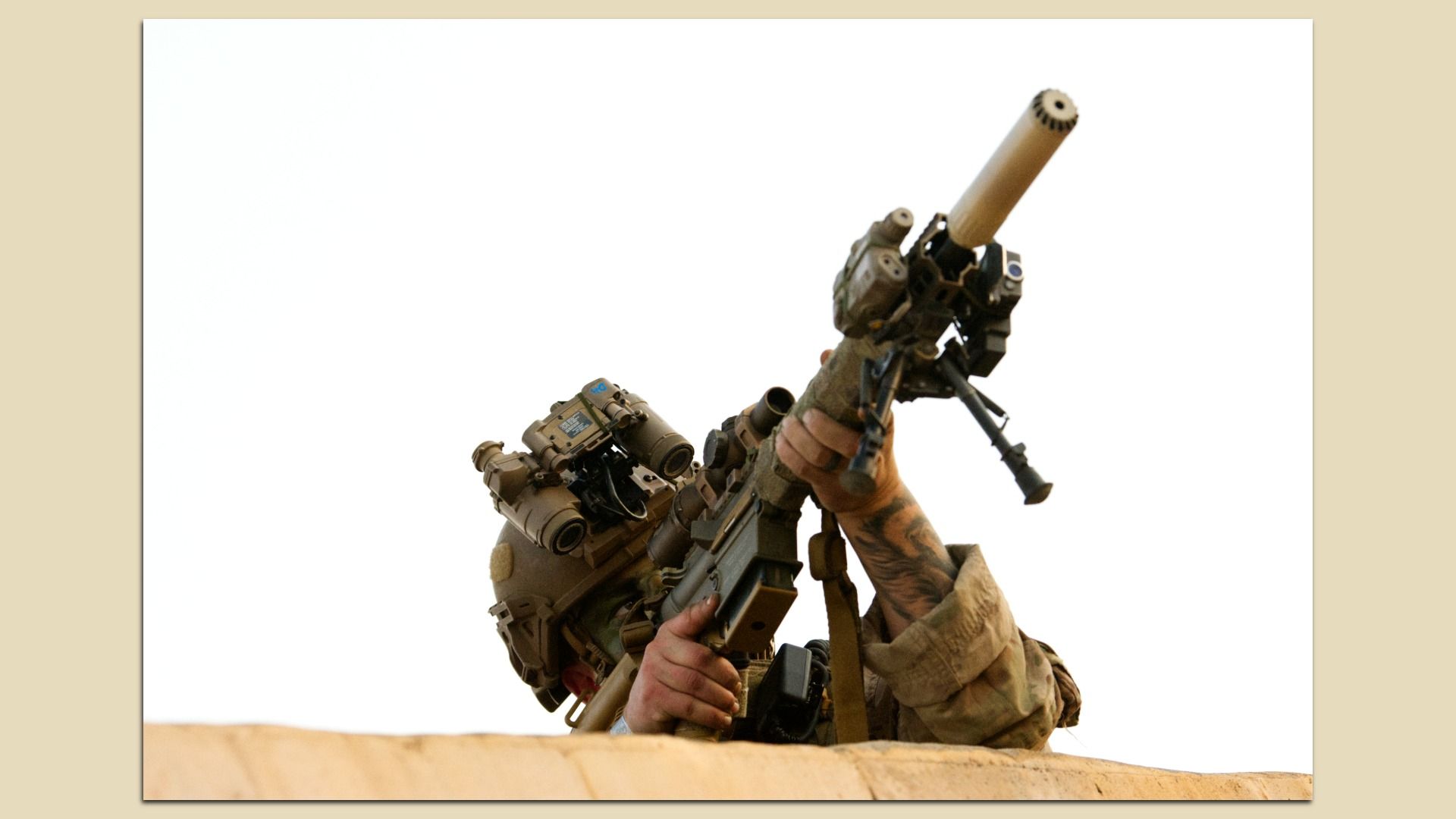 Desert-camouflaged soldier aiming a large rifle with a mounted scope and attachments, raised above a sandy ledge against a bright white sky.