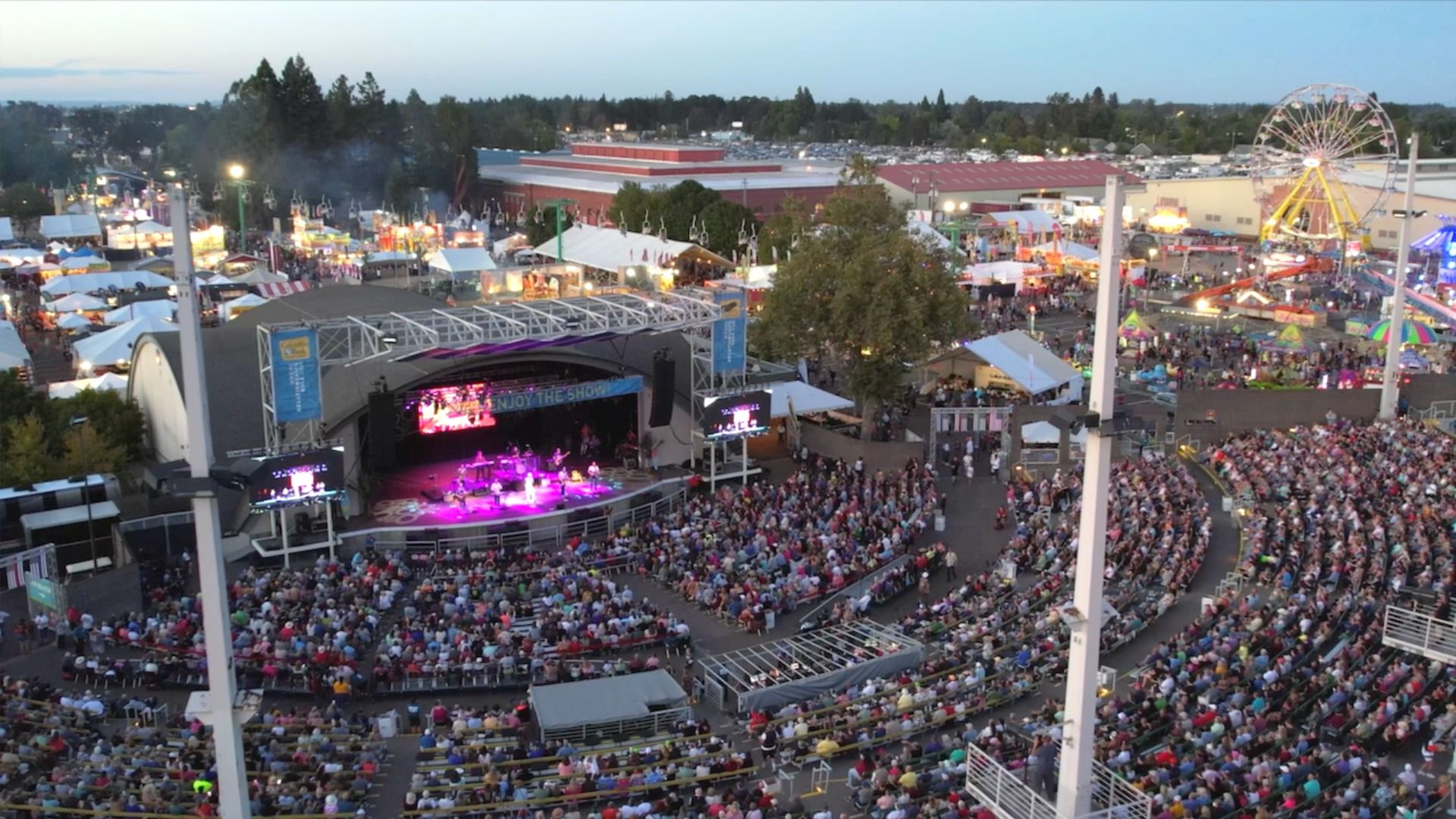 A large outdoor amphitheater filled with people watching a live concert on a lit stage at dusk, with a brightly lit fair featuring a Ferris wheel and tents in the background.