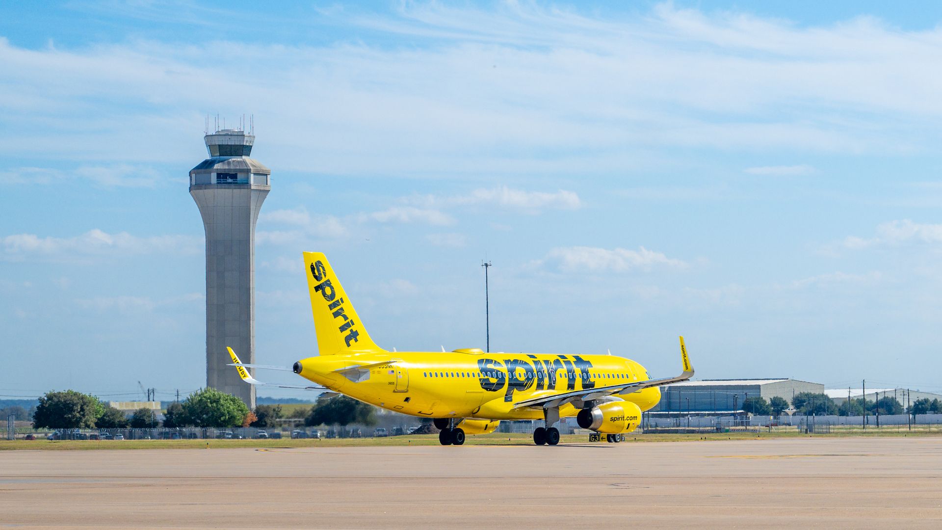 Bright yellow Spirit Airlines jet on the taxiway beside a tall airport control tower under a clear blue sky with wispy clouds.